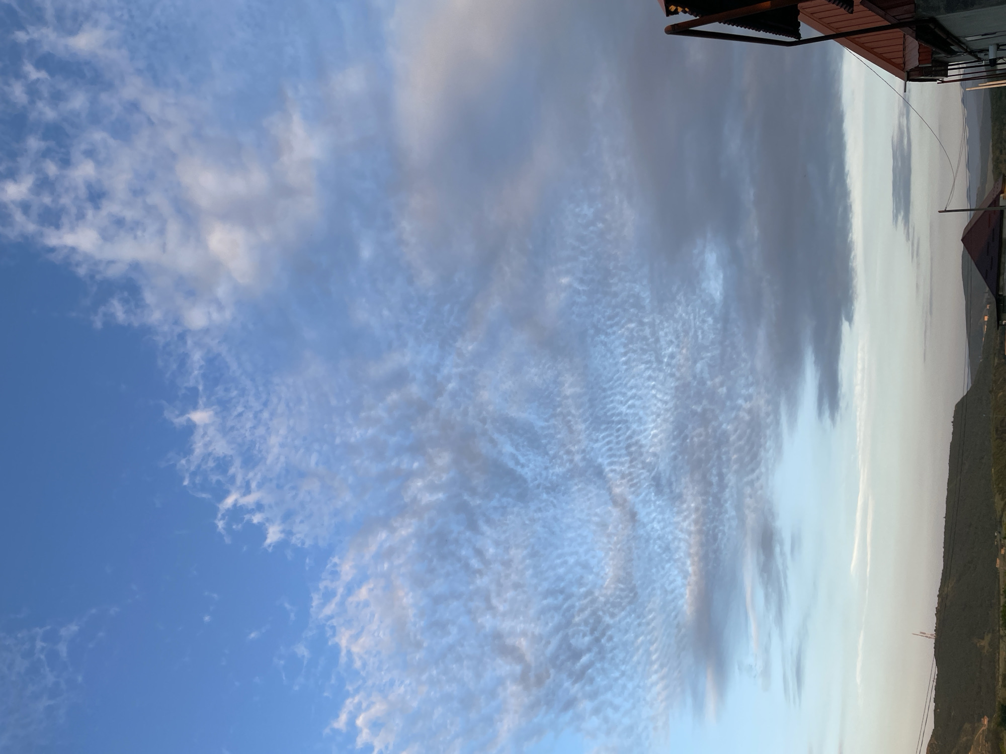 This media file captures a wide view of a sky with dramatic cloud formations above a mountainous landscape. The clouds range from thin, wispy cirrus to denser, darker formations, suggesting changing weather or atmospheric conditions. The sky transitions from a vibrant blue on the left to a lighter, hazy grey towards the right, where the sun is likely setting or has recently risen.
In the foreground, the tops of buildings with reddish-brown roofs are visible, indicating a human settlement nestled amongst the hills. One building has an overhang supported by metal poles, adding an architectural detail. The landscape features rolling hills and mountains, sparsely vegetated, with power lines and possibly a radio tower visible in the distance. The overall scene evokes a sense of natural beauty and the vastness of the sky.
The lighting suggests it is either early morning or late evening, with the sun low on the horizon. The weather appears partly cloudy, with some areas of clear blue sky and others obscured by cloud cover. There is no visible text in the image. The media file is oriented vertically, as if taken looking upwards at an angle. Given the location context of Buynaksk, Russia, the scene likely depicts a rural or semi-rural area within that region.