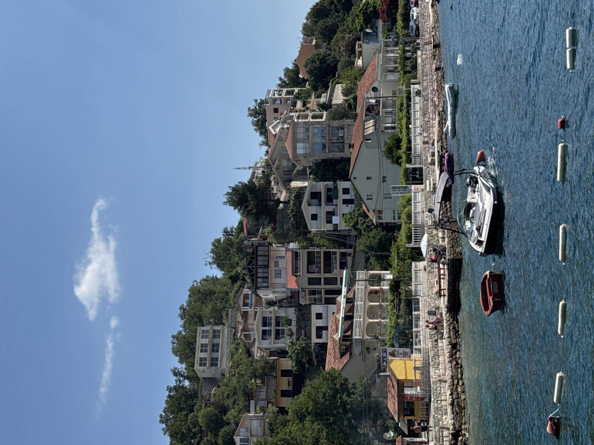 This image depicts a waterfront scene in a hilly coastal town under a clear blue sky. The foreground is dominated by the deep blue, slightly rippled water of a bay or sea. Several white buoys are lined up horizontally in the water, with a red buoy closer to the left edge. Two small boats are visible in the water: a red dinghy in the middle ground and a white and black motorboat with a canopy in the right middle ground, appearing to be in motion.
Along the shoreline, a stone embankment forms a walkway or promenade. Behind this, a row of buildings, varying in style and color, are situated on the sloping hillside. Some of these buildings are residences, characterized by balconies, windows, and terracotta roofs, while others appear to be smaller commercial establishments, including a yellow building with a sign and a canopy. People are sparsely visible: a man stands near the yellow building on the left, and a couple sits on a stone bench on the promenade. Further up the hill, numerous houses and apartment buildings are interspersed with lush green trees and foliage, climbing up the steep terrain. The architecture ranges from modern to older styles, with different colored facades and roof types. The overall impression is of a picturesque Mediterranean or similar coastal settlement bathed in bright sunlight.