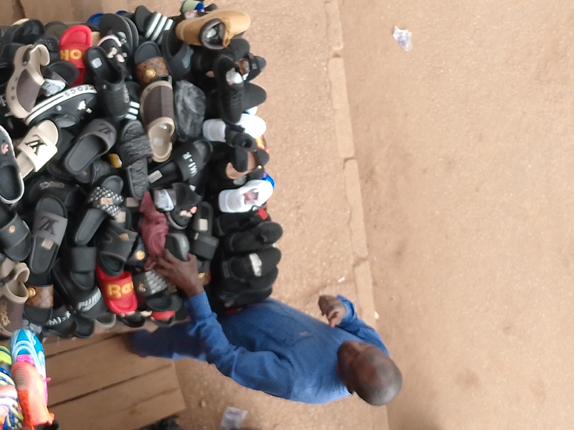 In Bukuru, Nigeria, a person in a blue long-sleeved shirt is standing in front of a large display of footwear. The footwear, primarily sandals and flip-flops, is stacked in a wooden cart. The display includes various colors, brands, and styles. The ground is a light brown, sandy surface.