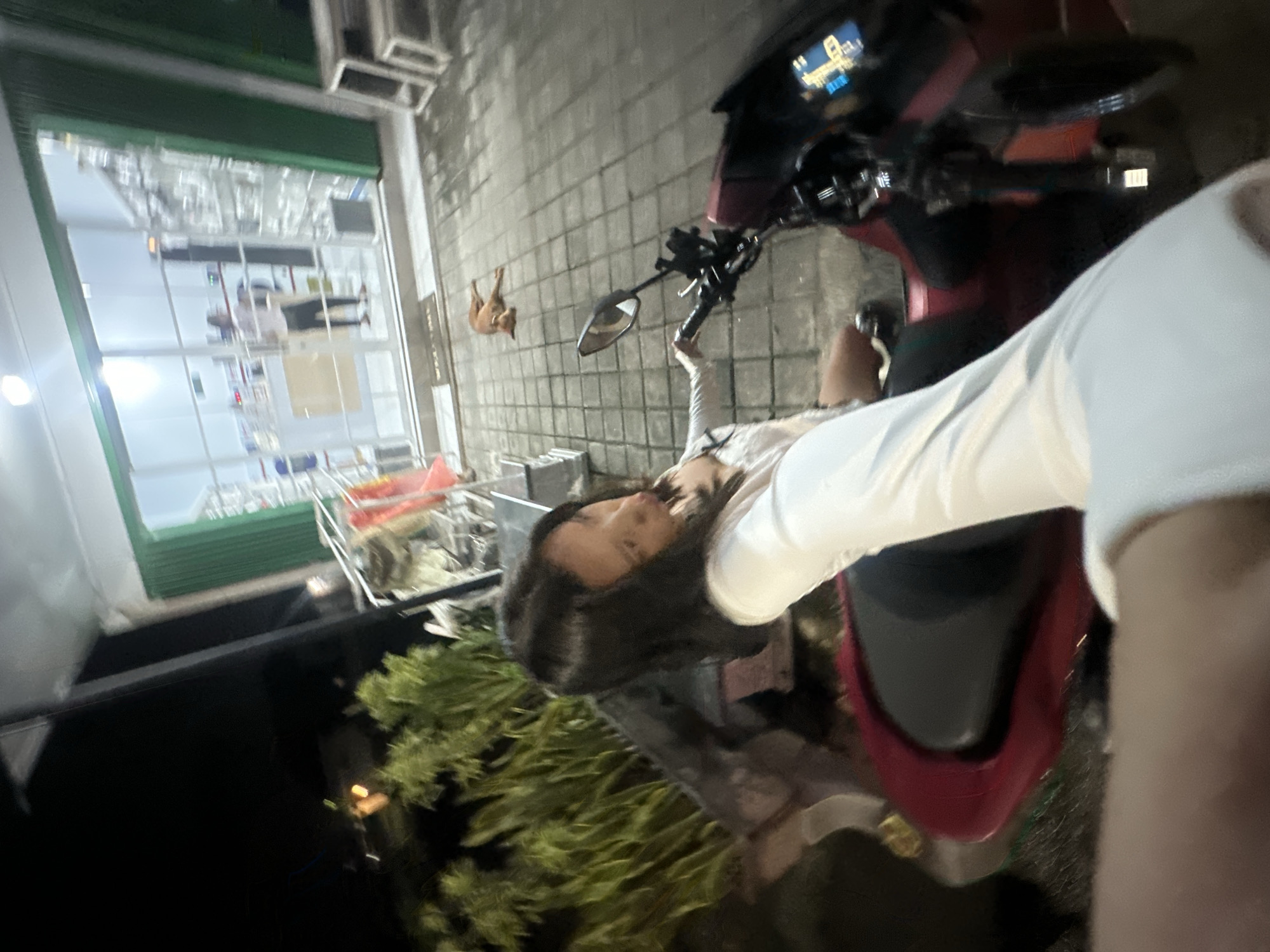 A person sits on a red scooter parked on a tiled surface in what appears to be North Kuta, Indonesia. A dog is nearby. Behind the scooter and person is a building with a large glass window and metal frame. The surrounding environment includes vegetation. The scene appears to be taken at night or in low light conditions.
