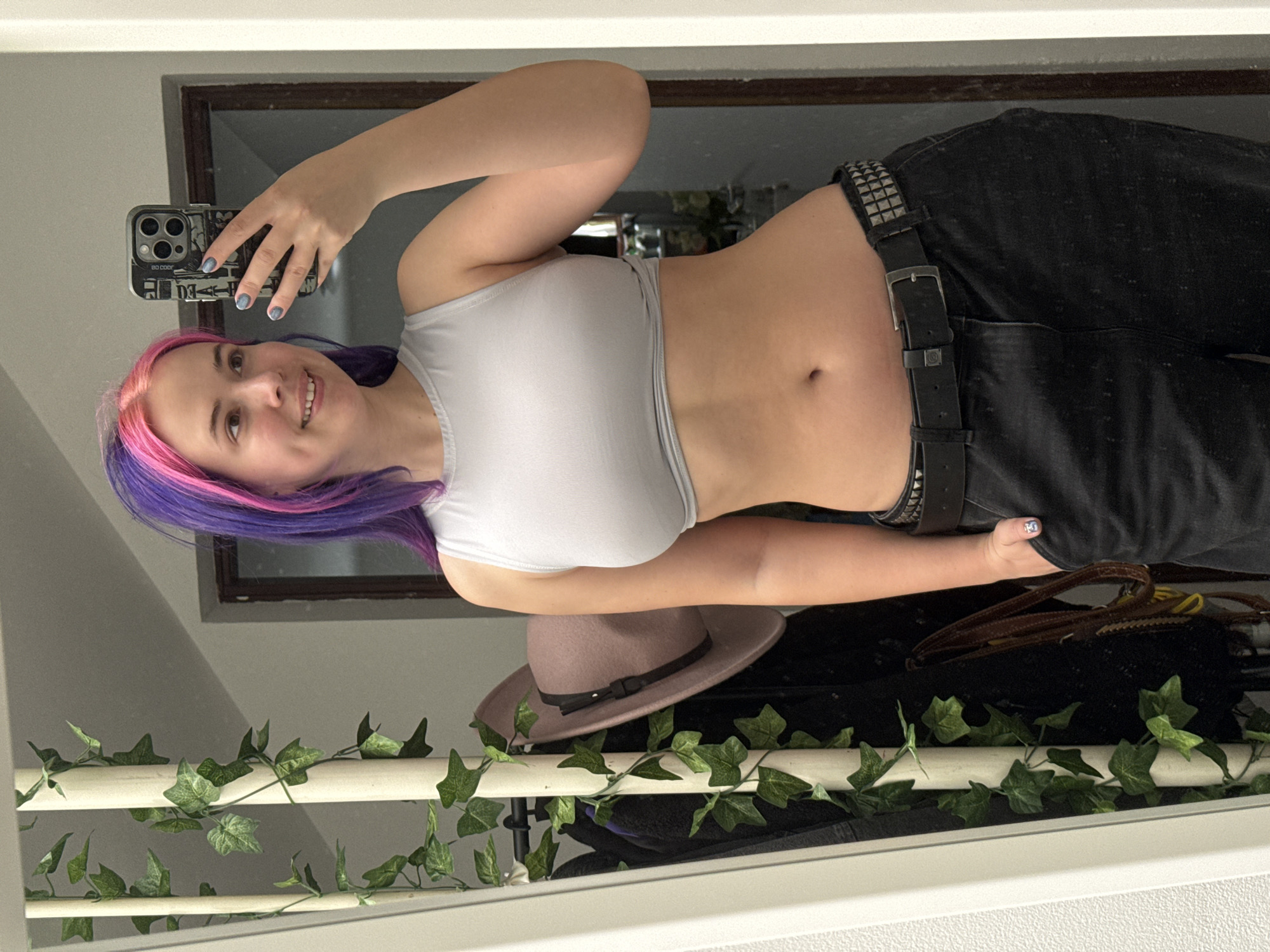A woman is taking a selfie in a mirror. She has pink and purple hair and is wearing a gray crop top and black pants with a studded belt. A hat and other items are on a shelf or surface behind her. Decorative ivy is wrapped around a white pole or railing in the foreground. The reflection shows a window and part of a room. The location is unknown, and there is no information to suggest it is in the City of Zagreb, Croatia.