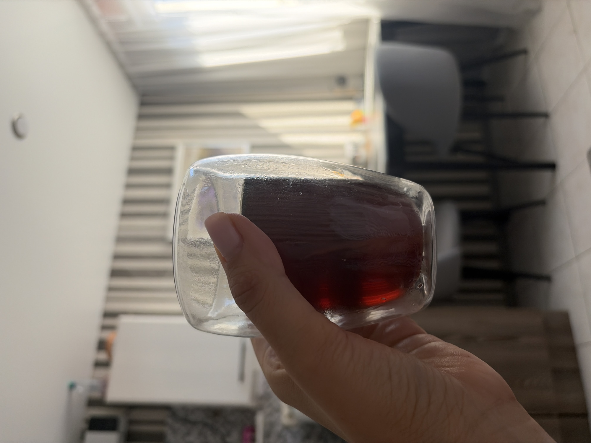 A hand holds a clear double-walled glass filled with a dark red liquid, possibly tea or juice. The glass is positioned in the foreground, slightly to the left of center, obscuring a portion of the background. The background depicts an interior scene, likely a kitchen or dining area. A table with two chairs is visible, with light filtering through sheer white curtains on the right. The wall behind the table has vertical stripes. The overall lighting suggests it is daytime, with sunlight casting shadows and highlights. The image appears to be a casual snapshot taken indoors. There is no indication of the city of Sochi, Russia, or any specific location details beyond the interior setting.