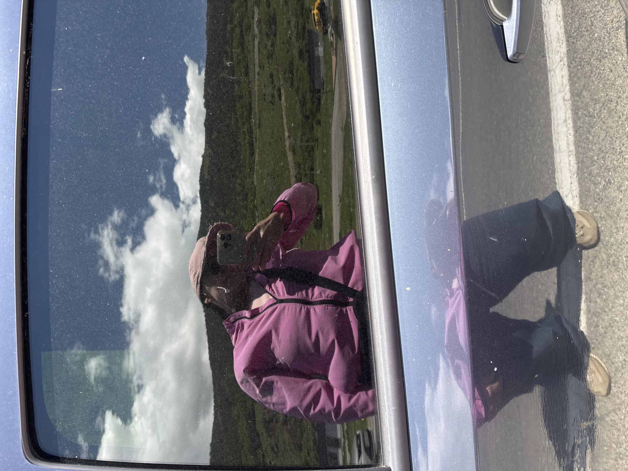 A reflection in a car window shows a person wearing a pink jacket and a pink hat, holding a smartphone to take a selfie. The reflection also captures a cloudy blue sky and a green, hilly landscape. Below the car window, the reflection on the car's surface shows the person's lower body, wearing dark pants and light-colored shoes, with the texture of the car's paint visible. In the background reflection on the window, a yellow vehicle is visible on a road. The surrounding environment appears to be a mountainous area with green slopes, consistent with a location like Верхне-Тебердинское сельское поселение, Russia.