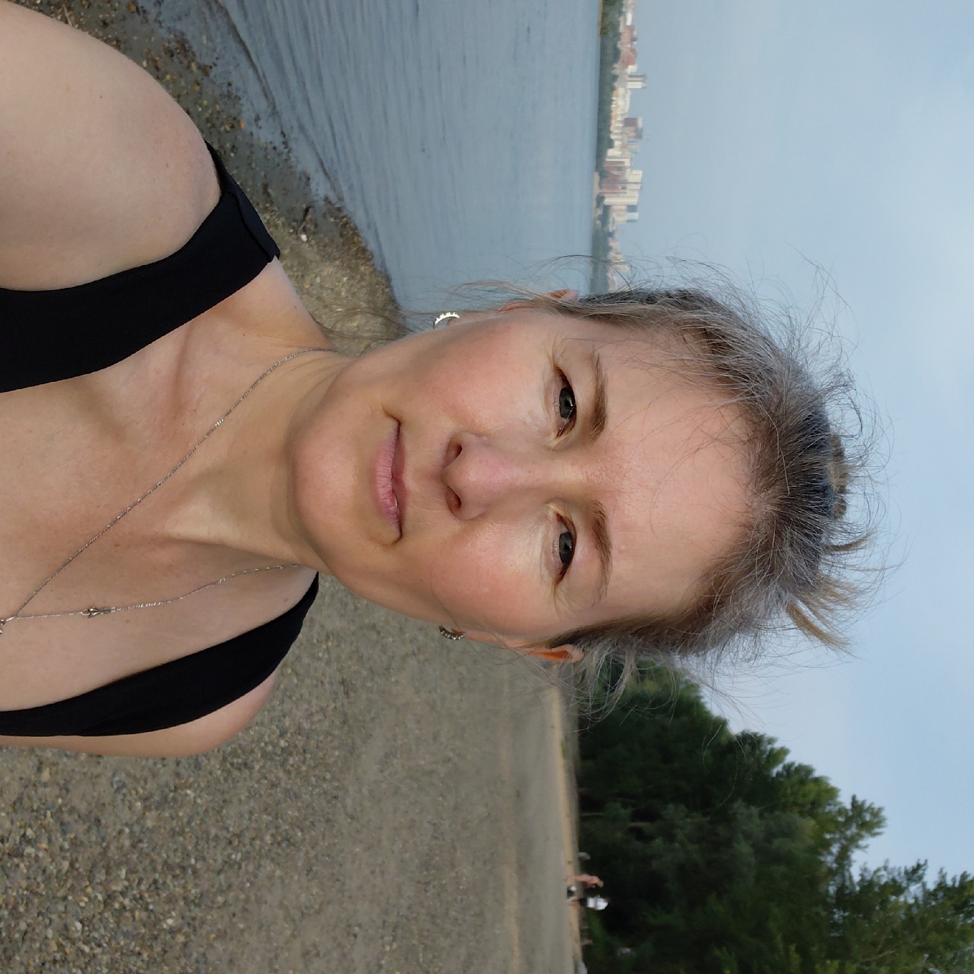This is a medium shot, eye-level, outdoor selfie taken on a pebble beach in Novosibirsk, Russia. The subject is a woman with graying blonde hair pulled up messily, fair skin, and light blue eyes. She is wearing a black sleeveless top and a silver necklace. She has small silver hoop earrings. She is facing the camera and smiling slightly.
Behind her, to the left, is a large body of water, likely the Ob River, with a city skyline in the distance featuring several multi-story buildings. To the right of the frame is a sandy beach dotted with pebbles and some sparse vegetation. Further in the background on the beach, two people are visible; one appears to be wearing a pink garment and another is in white, with a dark object on the ground between them. The overall lighting suggests it is late afternoon or early evening. The trees in the background are lush and green, indicating a summer or early autumn setting.