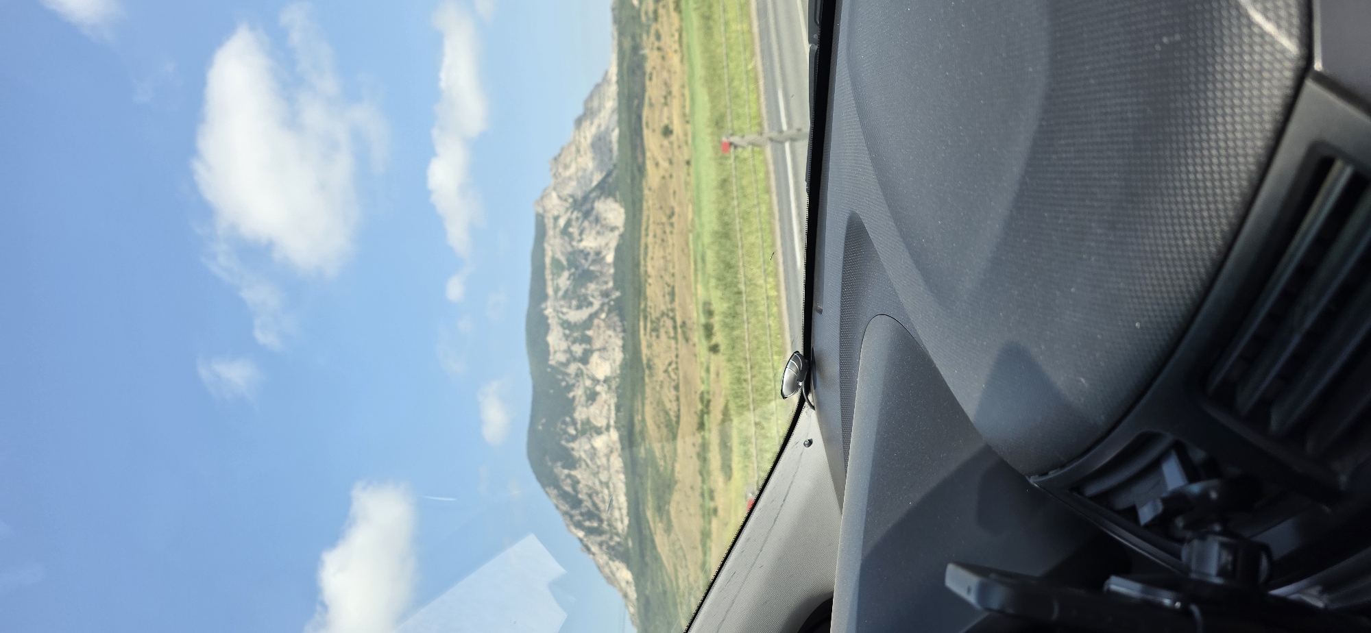 The image shows a view from inside a car, looking out through the windshield at a large, rocky mountain under a blue sky with white clouds. In the foreground, the car's dashboard is visible, with a small black device attached to the windshield. The mountain is covered in green vegetation at its base, transitioning to rocky, lighter-colored terrain towards its summit. A road and a green field are visible between the car and the mountain. There are no people visible in the image. The location is identified as Mineralnye Vody, Russia.