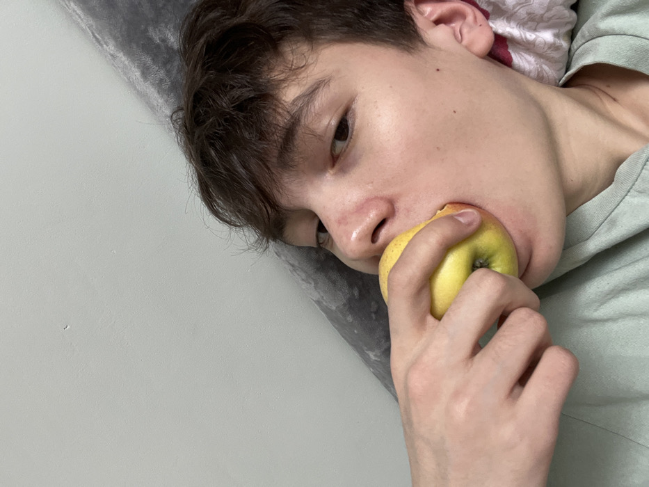 A close-up image depicts a young individual holding a yellow apple to their mouth with their right hand, appearing to be in the process of biting or consuming it. The individual has short, dark brown hair and is looking directly at the camera. A light green garment is visible on the subject. In the background, a dark grey textured fabric, consistent with a pillow or blanket, is visible behind the subject's head, contrasting with a plain light grey wall in the upper left of the frame. The scene is located in Novyi Nalim, Russia.