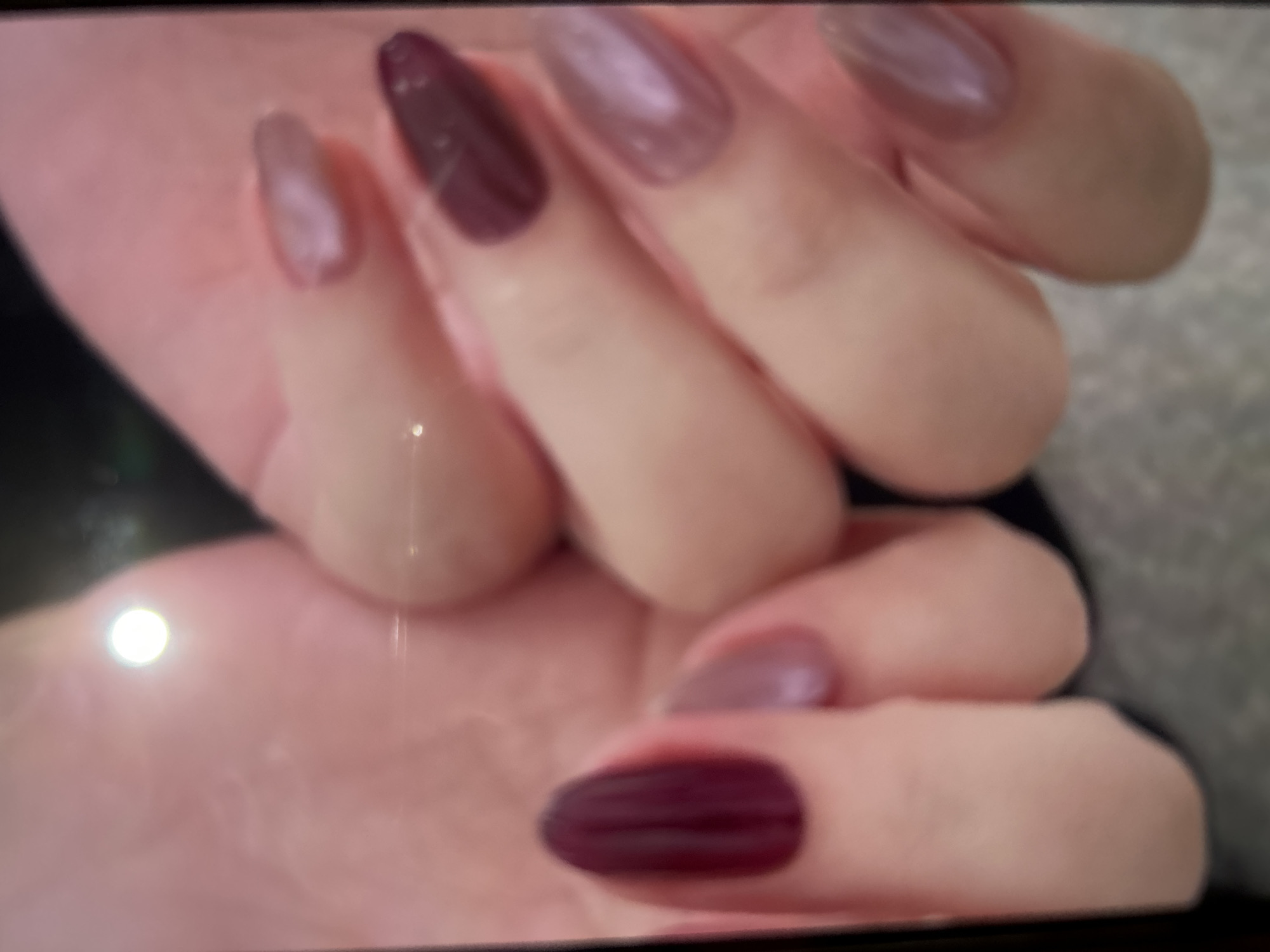 In Depok, Indonesia, a close-up image shows a person's hands with manicured nails. The nails are painted in various shades of deep red, muted purple, and a shimmery pinkish-beige. The background is out of focus and appears to be a textured surface, possibly a table or floor. A bright light source is reflected on the skin of the left hand.