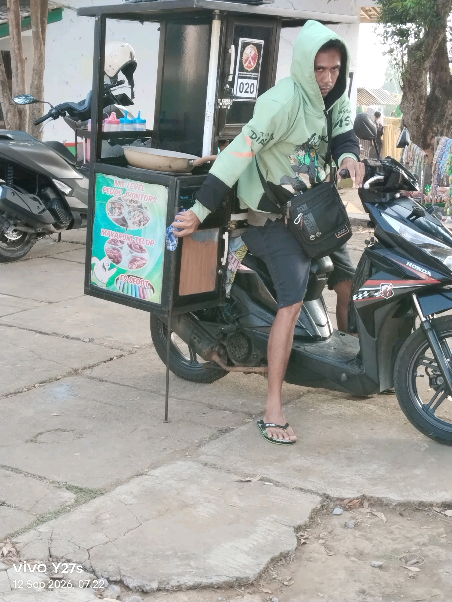 A medium shot captures a man standing next to a mobile food cart attached to a motorcycle, likely in Tamansari, Indonesia. The man, wearing a light green hooded sweatshirt and dark shorts, has a black satchel slung across his chest. He is looking towards the camera with a neutral expression. The food cart displays colorful images of food items and text, including "MIE LEVEL PEDAS JHONTORS," "MAKARONI TELOR," and "ES SEDOT." A white helmet is secured on the cart. The motorcycle is black and red with "HONDA" branding. The scene takes place on a paved outdoor area, possibly a roadside or market, with a building in the background and trees visible. The ground is concrete with some cracks and vegetation. The image is dated September 12, 2026.