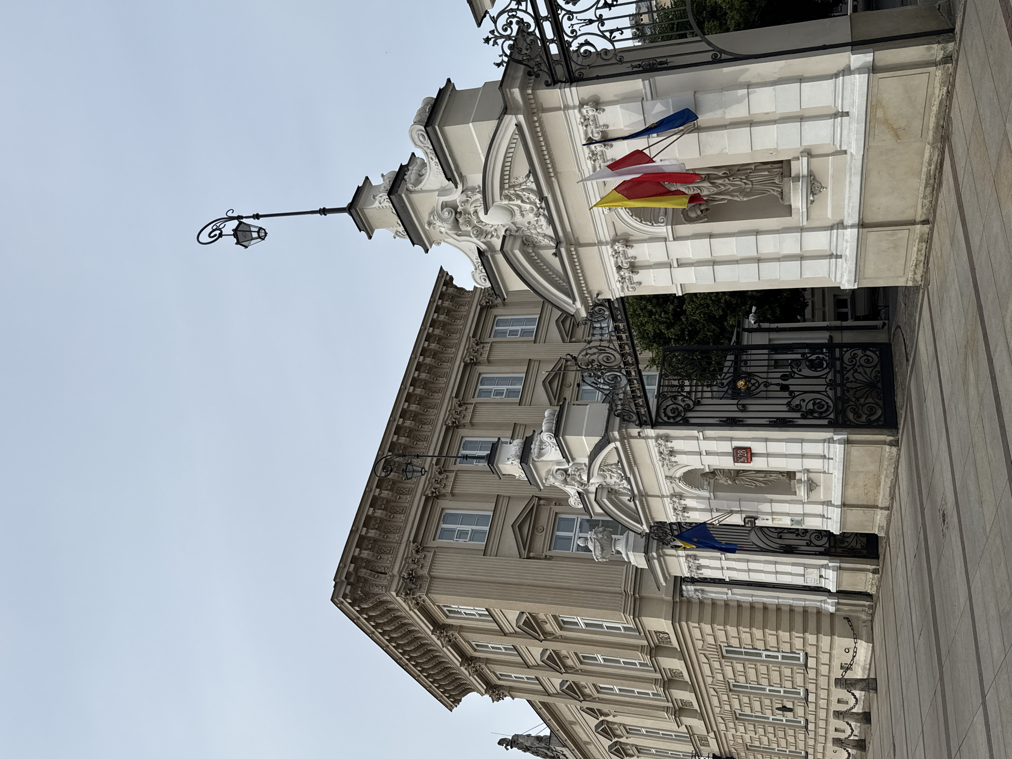 The image shows the exterior of a grand, ornate building with neoclassical architectural elements. The building is adorned with intricate carvings, columns, and decorative stonework. In the foreground, there is a wrought-iron gate and a stone pathway. Several flags are visible, including what appear to be the flags of Poland and the European Union, as well as smaller flags with red, white, and yellow colors. A lamppost with an old-fashioned design is also present. The sky is clear and bright, suggesting daylight. The overall impression is one of historical significance and architectural beauty. A red plaque with white numbers "2628" is visible on the building's facade. The orientation of the image is rotated 90 degrees clockwise.