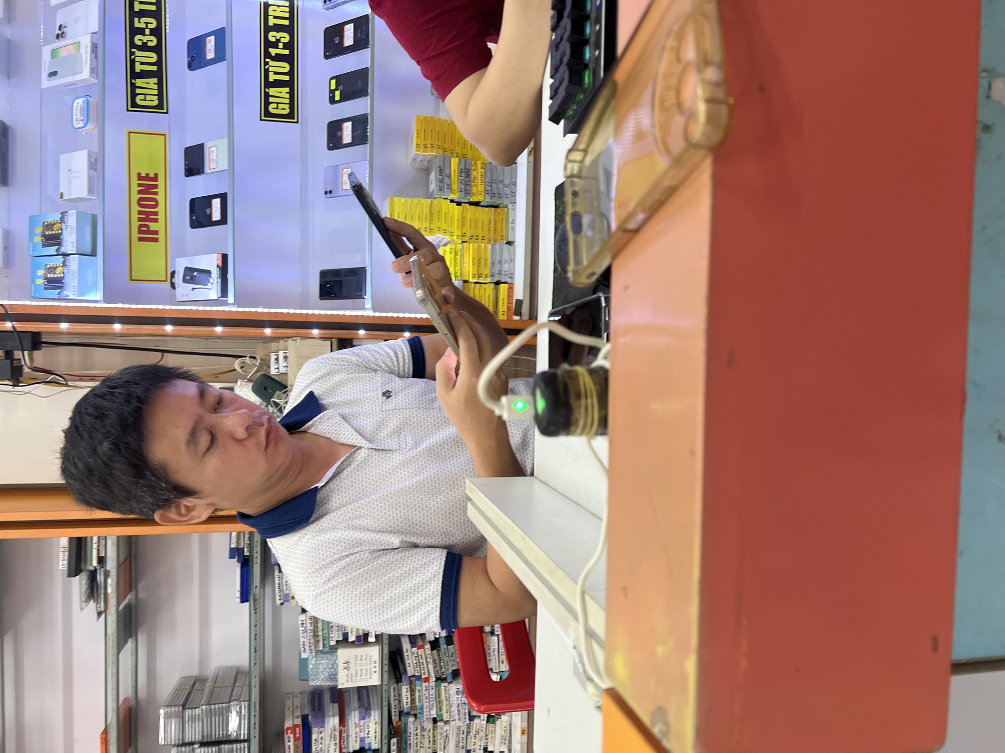The image depicts the interior of an electronics store, likely specializing in mobile phones, located in Đà Nẵng, Vietnam. A man of East Asian descent, wearing a white polo shirt with a blue collar, is seated behind a counter and appears to be examining a smartphone. Shelves filled with various phone cases and mobile phone packaging are visible in the background, along with illuminated signage advertising iPhones. The counter is cluttered with electronic accessories, including charging cables and a green charging indicator light. Another person's arm, wearing a red shirt, is visible on the right side of the image, also near the counter. The overall environment suggests a retail setting focused on mobile technology.