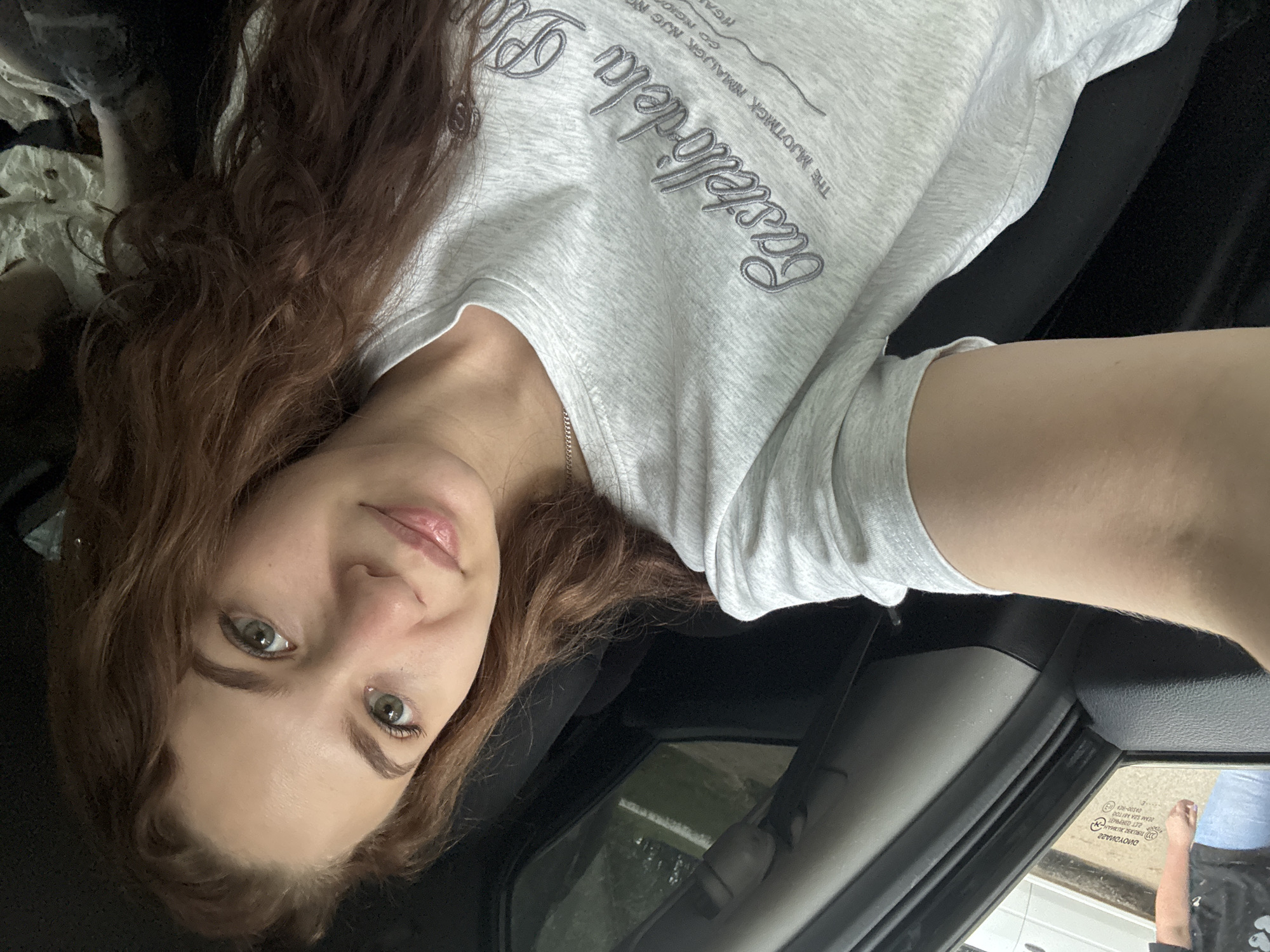 A woman with long, wavy auburn hair and light green eyes is taking a selfie from inside a car. She is wearing a light gray t-shirt with the words "Bastellídela Pla" and other text in smaller font. The interior of the car is visible, including the dashboard and door frame. A rearview mirror with stickers is present. The outside environment is blurred but suggests an outdoor setting with light-colored surfaces and greenery. The location, Chaikovsky, Russia, cannot be confirmed from visual elements in the picture alone.