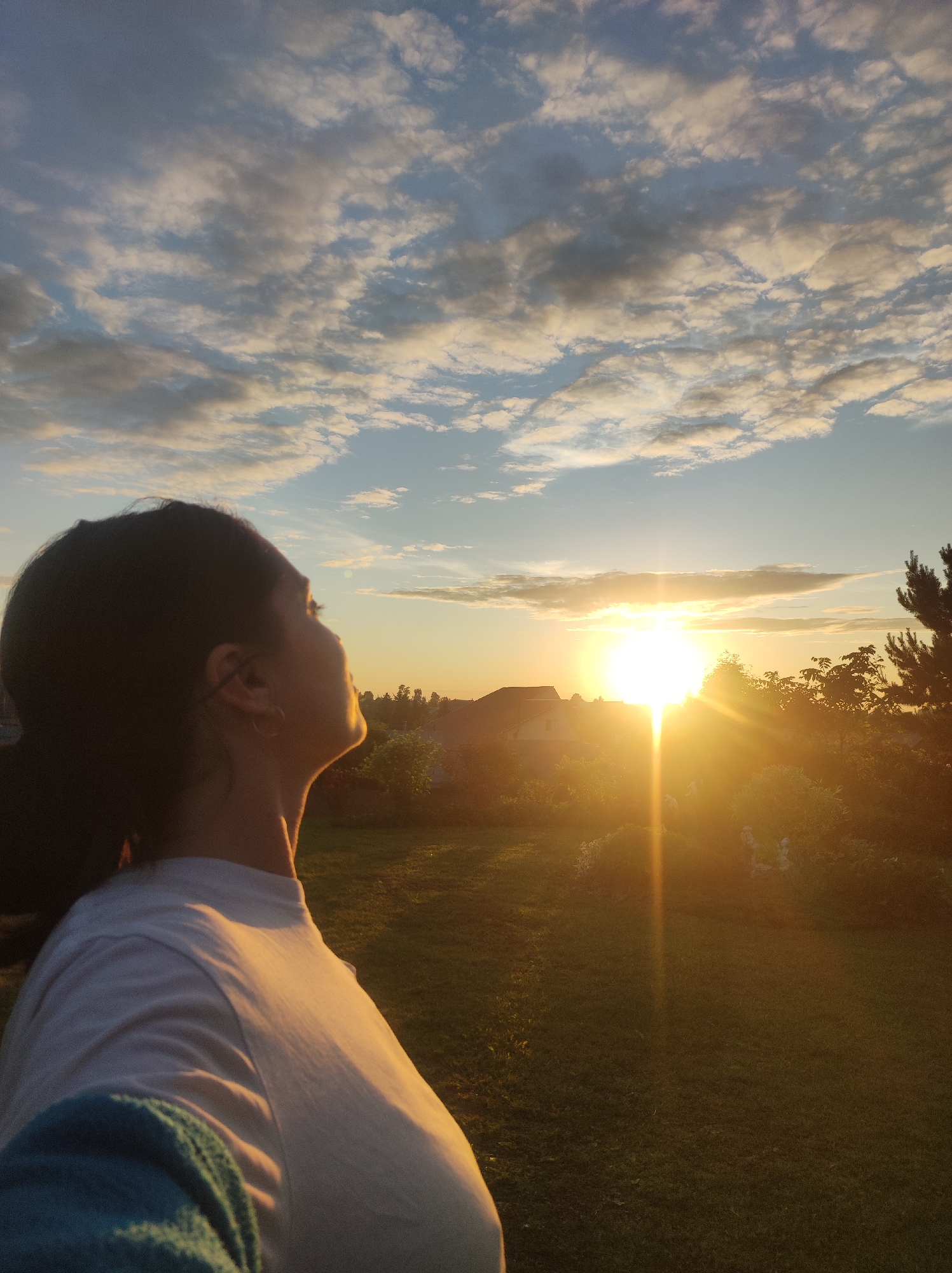 An outdoor scene captured in Фоки, Russia, features a female subject in profile, facing right and looking upwards towards a low sun. The sun, positioned on the right side of the mid-ground, emits a strong, vertical lens flare, indicating either sunrise or sunset. The sky above is partially cloudy, featuring scattered white and light grey clouds against a pale blue background, with some undersides showing darker tones. The female subject has dark hair pulled back and is wearing a white short-sleeved top, with a portion of a blue or teal garment visible on her left shoulder. Her face and neck are primarily in silhouette due to the backlighting, though a faint warm glow outlines her profile and a small hoop earring is visible on her ear. The background beyond the sun includes silhouetted trees, bushes, and several distant residential structures. The foreground consists of green grass, extending towards the mid-ground where additional foliage is present.