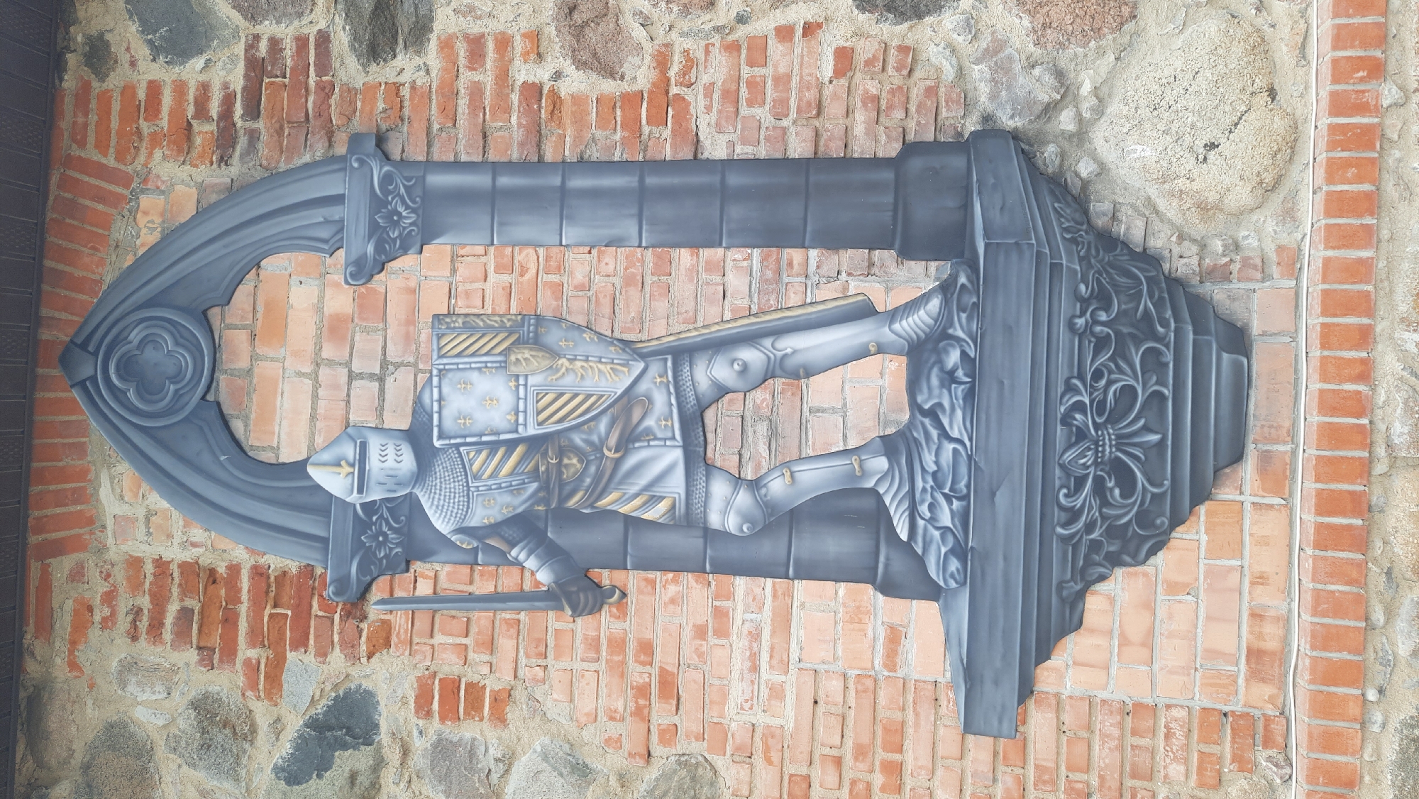 This is a photograph of a mural on a brick and stone wall, depicting a knight in full armor. The knight stands on a decorative stone pedestal, holding a sword in its right hand and a shield in its left. The mural is framed by a gothic-style archway with columns. The brickwork of the wall is visible behind and around the mural, with varying shades of red and orange bricks, interspersed with rough-hewn stones. The mural itself is rendered in shades of gray and metallic gold, with the knight's armor detailed with intricate patterns. The setting appears to be an exterior wall, possibly part of a historical building or a decorative element in the city of Жодзішкаўскі сельскі Савет, Belarus.