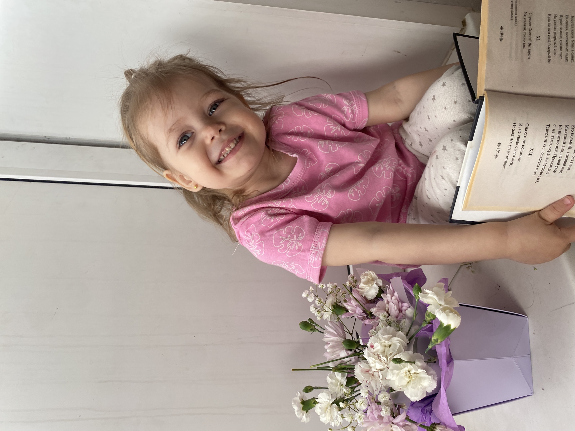 A young child with light brown hair pulled back in a ponytail is smiling brightly at the camera in this portrait-oriented image. The child, likely a girl, has blue eyes and is wearing a pink long-sleeved shirt with white leaf patterns and light-colored pants with small star designs. A gold stud earring is visible in her left ear. She is positioned on what appears to be a light-colored surface, possibly a window sill or bed, in an indoor setting.
In her hands, the child is holding an open book with visible text in Russian. The pages contain poetic verses, with chapter numbers "XL," "XLI," and "XLII" clearly visible, along with page numbers "194" and "195." The text mentions "Онегин," suggesting the book is likely Alexander Pushkin's "Eugene Onegin." To the right and below the child, a bouquet of light-colored flowers, primarily white and light purple chrysanthemums and carnations, is arranged in a purple box or wrapper.
The lighting is bright and even, indicating daytime. The child's cheerful expression and relaxed pose suggest a happy and comfortable moment. The scene takes place in Chaikovsky, Russia.