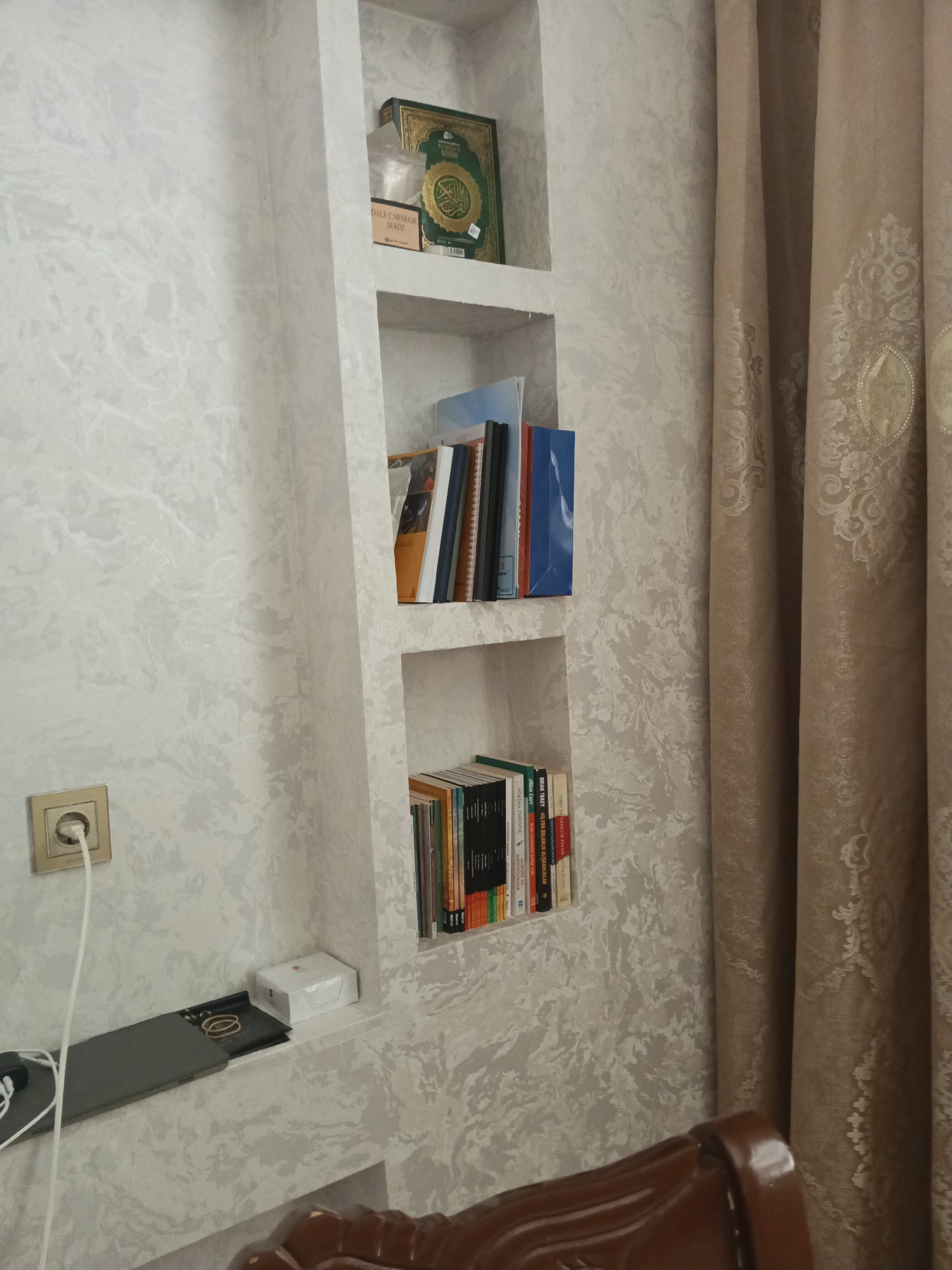 A framed green Quran is placed on the top shelf of a built-in wall niche, next to a box of books. The second shelf holds a collection of books, some with colorful spines, stacked vertically. The bottom shelf displays more books, also arranged vertically, alongside a white box and a flat dark gray object. To the left of the niche, a gold-colored electrical outlet with a white cord plugged in is visible on a textured light-colored wall. A brown upholstered chair is partially visible in the lower right corner of the frame, and a beige patterned curtain hangs to the right of the niche. The scene is indoors, likely in a residential setting in Masazir, Azerbaijan.