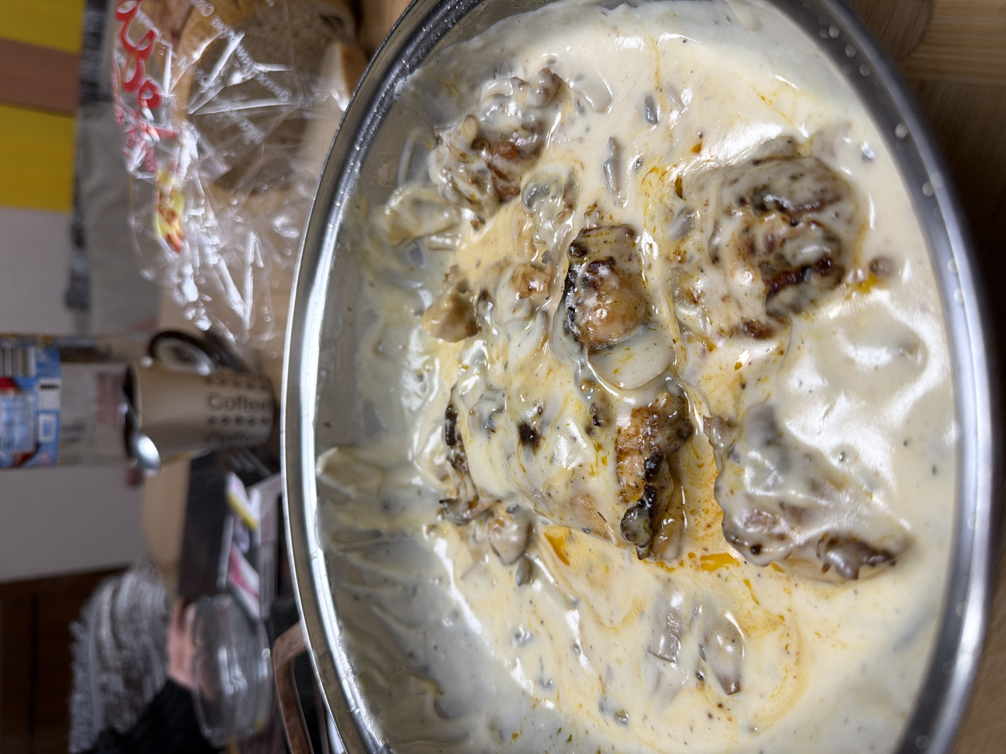 A top-down, close-up shot shows a stainless steel pot filled with a creamy sauce and browned meat pieces. The sauce is white-to-off-white with some yellowish oil slicks and small brown specks. The meat pieces are irregularly shaped and appear to be cooked, with some charring on the exterior. The pot is situated on a surface with miscellaneous objects in the background, including a plastic-wrapped loaf of bread, a coffee mug, and other items. The location in the city of Panagyurishte, Bulgaria cannot be determined from the visual data provided.