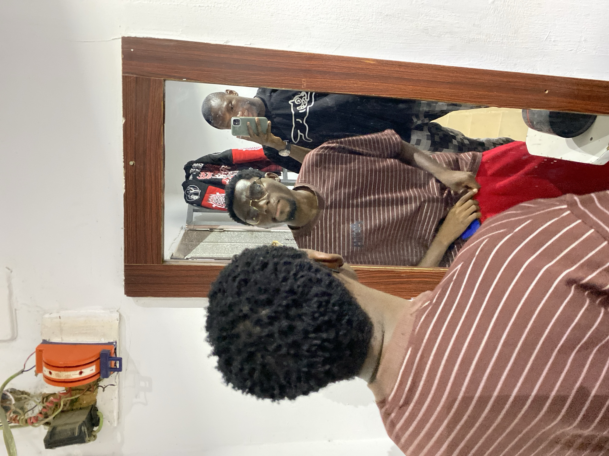 This image depicts an indoor scene featuring two men and a mirror. In the foreground, the back of a man with short, curly dark hair is visible. He is wearing a brown and white striped shirt and appears to be looking into the mirror.
Reflected in the mirror, framed in wood, is another man taking a selfie with a smartphone held in his hand. This man has short dark hair, a light beard, and wears glasses. He is dressed in a black long-sleeved shirt featuring a distinct white graphic of a cartoon-like character, possibly a bear or cat, with unreadable text beneath it. He is also wearing light-colored trousers or shorts.
Within the mirror's reflection, behind the man in the black shirt, various items are visible including red fabric and a black and red patterned garment or bag. A partial view of an interior space, possibly with a window or vent, is also reflected.
To the left of the mirror, mounted on the white wall, is an orange and blue electrical component with exposed wires, possibly an old switch or part of an electrical installation. The wall around this device shows some wear and tear. The scene suggests a casual indoor setting. No specific time of day or weather conditions are discernible from the image.