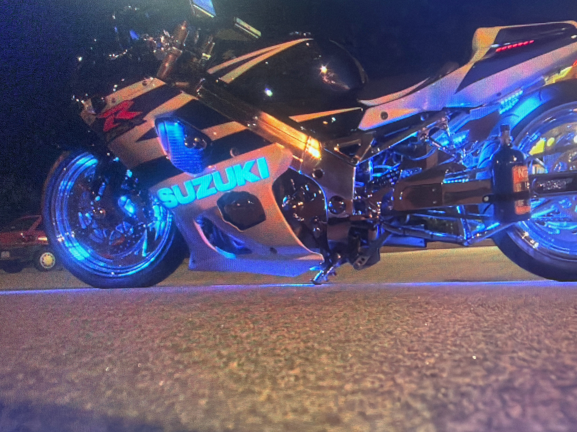 The image shows a Suzuki motorcycle illuminated by blue and orange lights, parked on an asphalt surface at night. The motorcycle is positioned at a low angle, with its front wheel and a portion of the engine visible. The "SUZUKI" logo is clearly displayed on the side of the motorcycle in a bright, cyan color. In the background, a red car is partially visible, and the environment appears to be a city street or parking area. The lighting suggests a custom or modified motorcycle, possibly with underglow lighting. The image does not provide any information that can be used to determine its location in the city of Zarafshan, Uzbekistan.