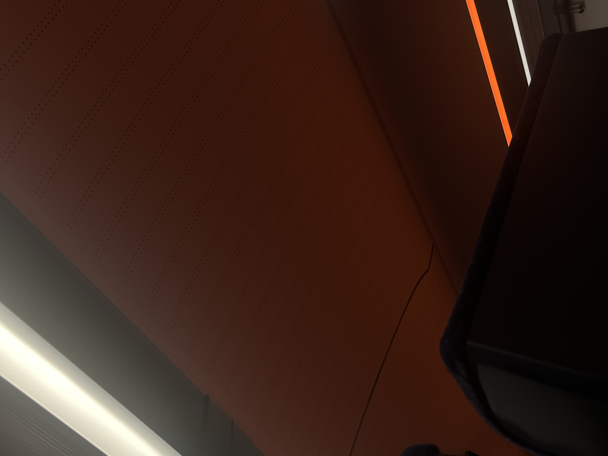 The image shows the interior of what appears to be a mode of transportation, possibly a train or bus, given the seating and overhead compartments. A dark-colored seat with a fabric covering is visible in the foreground, partially obscuring a dark electronic screen. Above the seat, a large, reddish-brown panel with a pattern of small, regularly spaced dots is angled upwards. To the left, a bright, white light strip runs diagonally across the top of the frame, casting a strong illumination. Below this, a section of a grey, corrugated surface is visible. Further to the right, a thin, vibrant orange light strip is present, running horizontally. The environment is dimly lit except for the light strips and the glow from the screen. The overall impression is one of modern, enclosed interior design. The location in Ilmenau, Germany, is not discernible from the visual information presented in the image.