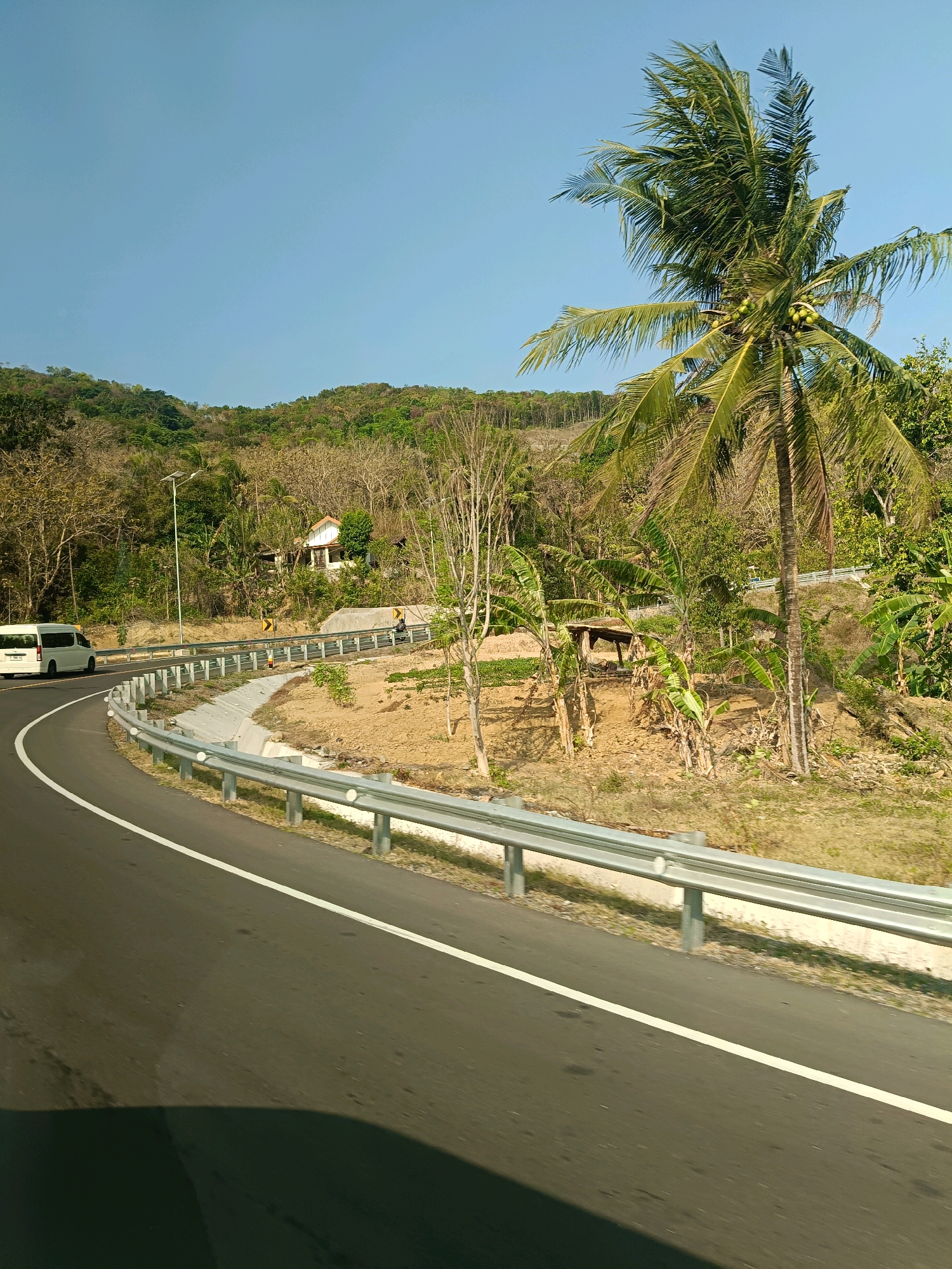 The image depicts a winding road with a guardrail, viewed from the perspective of a moving vehicle. A white van is visible in the distance on the left side of the road, traveling in the same direction. The landscape surrounding the road is hilly and covered with vegetation, including trees and some dry foliage. A tall coconut palm tree with coconuts is prominent on the right side of the frame. In the background, a small building with a white roof and light-colored walls is nestled among the trees on a hillside. The sky is clear and blue. The scene suggests a rural or semi-rural setting, possibly on the outskirts of Parangtritis, Indonesia, characterized by its coastal and hilly terrain.