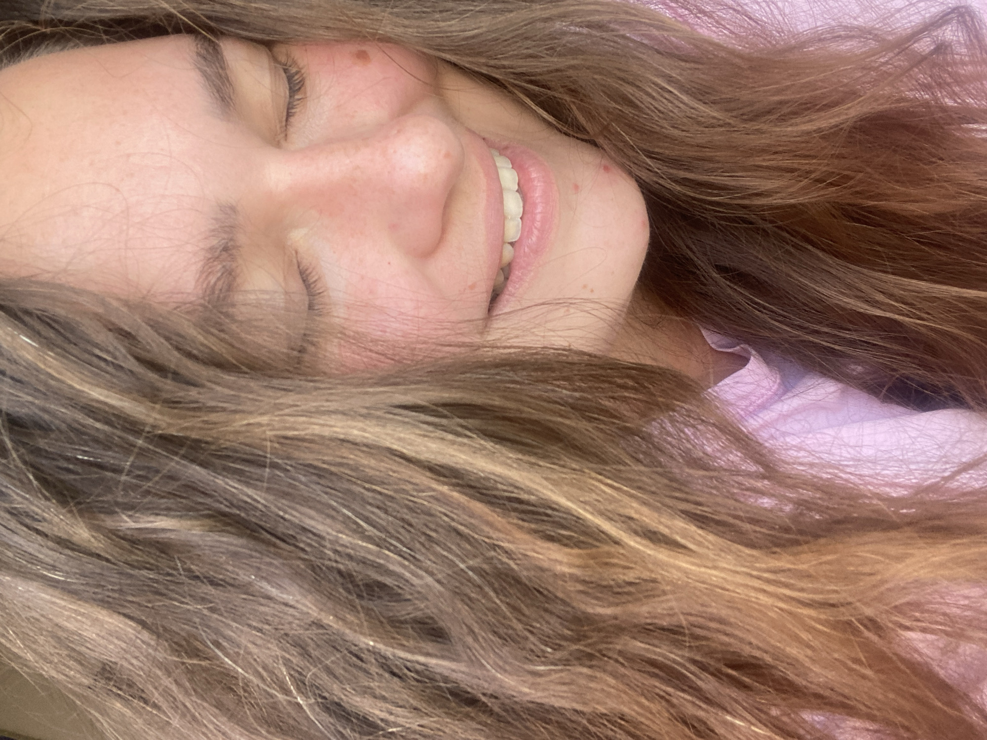 The image is a close-up, vertical, low-angle shot of a young woman's face, framed by her long, wavy, brown and blonde hair. Her eyes are closed, and she has a gentle smile, revealing white teeth. Her skin is fair with some light freckles and redness across her cheeks and nose, and a few small blemishes are visible on her chin and cheek. She is wearing a light pink shirt. The background consists of her hair and the pink fabric of her shirt. There is no discernible urban environment or the city of Perm, Russia.