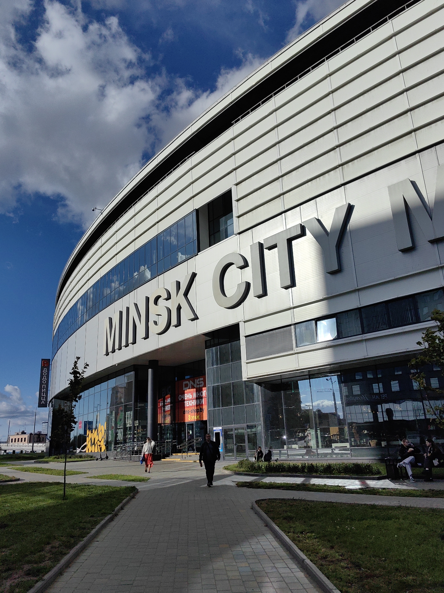 The image shows a wide shot of the Minsk City sports and entertainment complex in Minsk, Belarus, on a bright, partly cloudy day. The dominant subject is the modern, multi-story building with a curved facade, characterized by horizontal white panels and bands of reflective glass windows. Large, silver, three-dimensional letters spelling out "MINSK CITY" are prominently displayed on the upper section of the building.
In the foreground, a paved walkway leads towards the entrance of the complex, bordered by a green lawn and a small tree on the left. Several people are visible: a man in dark clothing walks away from the camera on the right side of the walkway, and further ahead, a woman in a red skirt and white top walks towards the entrance. A few individuals are seated on a bench on the far right, and others are entering or inside the building, which is largely visible through the large glass windows.
A vertical banner with Cyrillic text, possibly advertising "Dodo Pizza", is attached to a pole on the left side of the building. In the distance, across from the complex, are low-rise buildings with a darker brick facade. The sky is blue with scattered white cumulus clouds. The overall impression is of a modern urban environment under a dynamic sky.