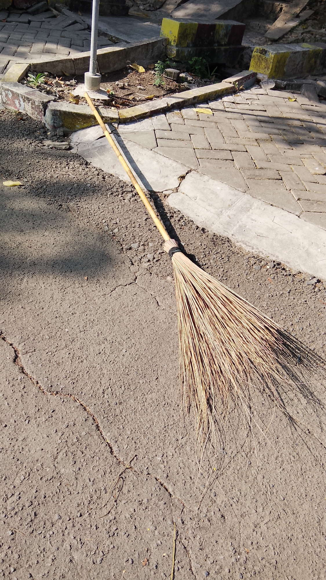 The image displays a traditional broom made of dried coconut leaves with a bamboo handle, resting on a cracked asphalt surface. The broom is positioned diagonally, with its bristles spread out and casting a shadow. In the background, there is a paved walkway made of interlocking concrete blocks, bordered by a concrete curb. Beyond the walkway, a small patch of dry soil with scattered leaves and a few sparse plants is visible, bordered by concrete structures. The scene appears to be outdoors, possibly in a public area within Tlogobendung, Indonesia, given the context of the request. The lighting suggests it is daytime, with sunlight creating distinct shadows. No individuals are present in the image.