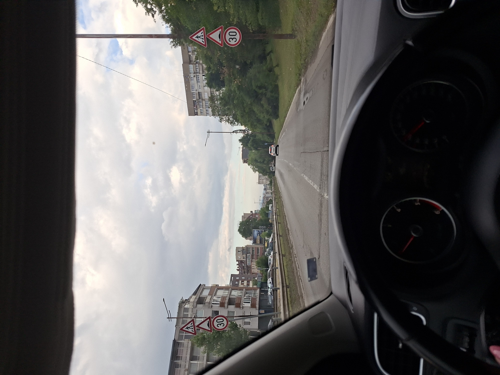 The media file shows a view from inside a car, looking forward and slightly to the left. The driver's side dashboard, including a speedometer, is visible on the right. The car appears to be driving on a road with a sidewalk and buildings on the left. There are several traffic signs visible: a triangular warning sign with a pedestrian crossing symbol, another triangular warning sign with a bumpy road symbol, and a circular sign indicating a speed limit of 30. The sky is cloudy, suggesting overcast weather. The buildings are apartment blocks with multiple stories and windows. Trees line the road between the sidewalk and the buildings, and also beyond the buildings. The scene appears to be an urban or suburban environment. The time of day is difficult to determine precisely due to the overcast sky, but it seems to be daytime. There are no people clearly visible in the image. The overall impression is of a routine drive through a residential or mixed-use area.