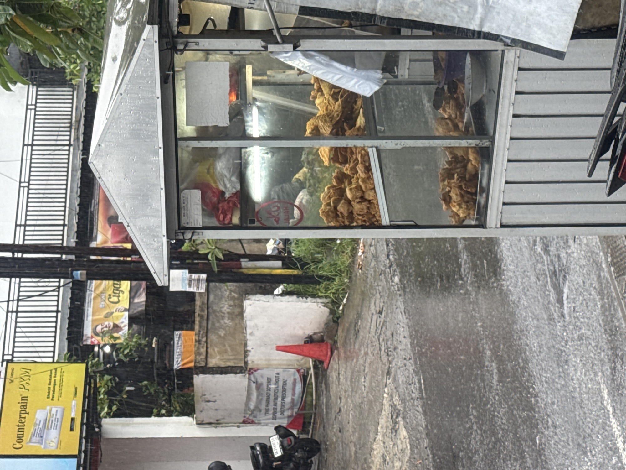 The image captures an outdoor street scene in Kuta Selatan, Indonesia, during what appears to be a rainy day, evidenced by the wet ground and visible water streaks. The photograph is oriented vertically, with the ground appearing on the right side.
A prominent metal food cart with glass display sections stands in the center-left. Inside the glass display, large quantities of golden-brown, irregularly shaped fried food, possibly fried chicken or a similar snack, are visible. Some plastic bags and other items are also stored within the cart. The cart has an angled, reflective metal roof.
In the background, on the left side of the image, there is a black metal gate with horizontal slats. Behind the cart, lush green foliage is visible, along with utility poles and tangled wires. Several signs and banners are attached to structures in the background. A large yellow sign on the far left advertises "Counterpain PXM" with text reading "Efektif Redaka" and "Peradangan Se." Another yellow banner in the background has partial text, possibly "Ciga."
Towards the bottom right, on the wet concrete ground, a bright red traffic cone is placed. Behind the cone, a white concrete block and part of a motorcycle are visible. A white sign affixed to the concrete block states: "THIS PHARMACY DO'S NOT DISPENSE ANTIBIOTICS WITHOUT A DOCTOR'S PRESCRIPTION." Inside the food cart, a small red label with "JAJAH" is discernible. No people are visible in the frame. The lighting suggests it is daytime.