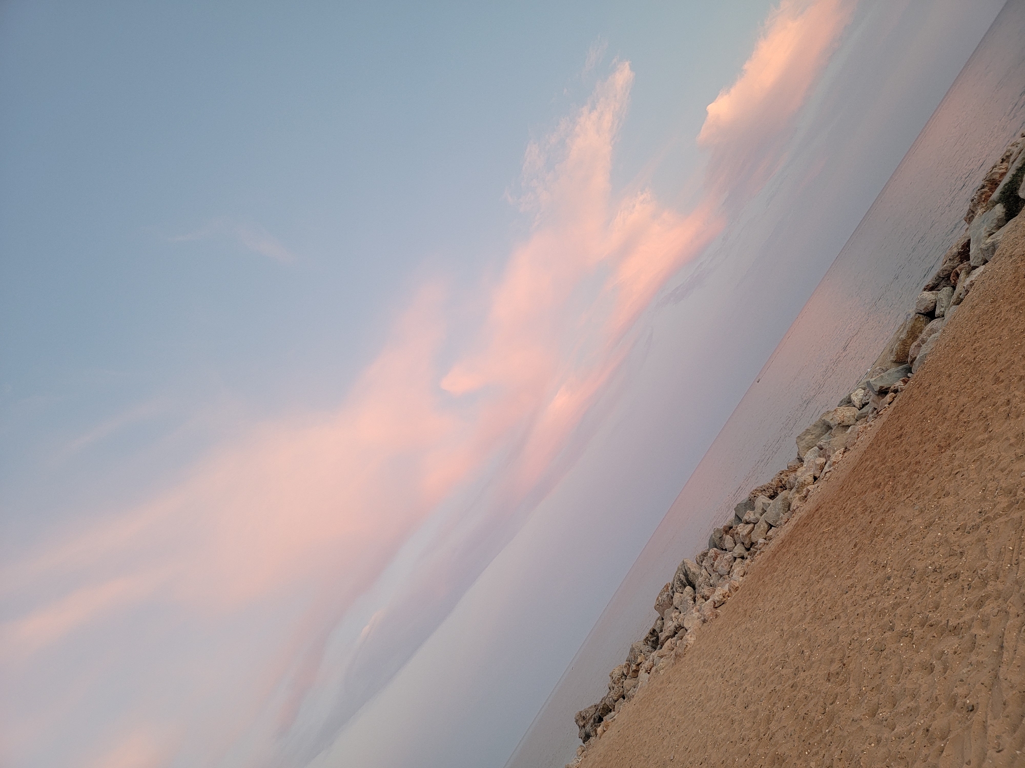 The image depicts a diagonal view of a sandy beach meeting the ocean, with a rock barrier along the shoreline. The sky above is a soft blue, transitioning to lighter hues near the horizon, and is adorned with wispy clouds tinged with pink, suggesting either dawn or dusk. The scene is static and serene. There are no people or animals visible. The location is identified as Quarteira, Portugal.