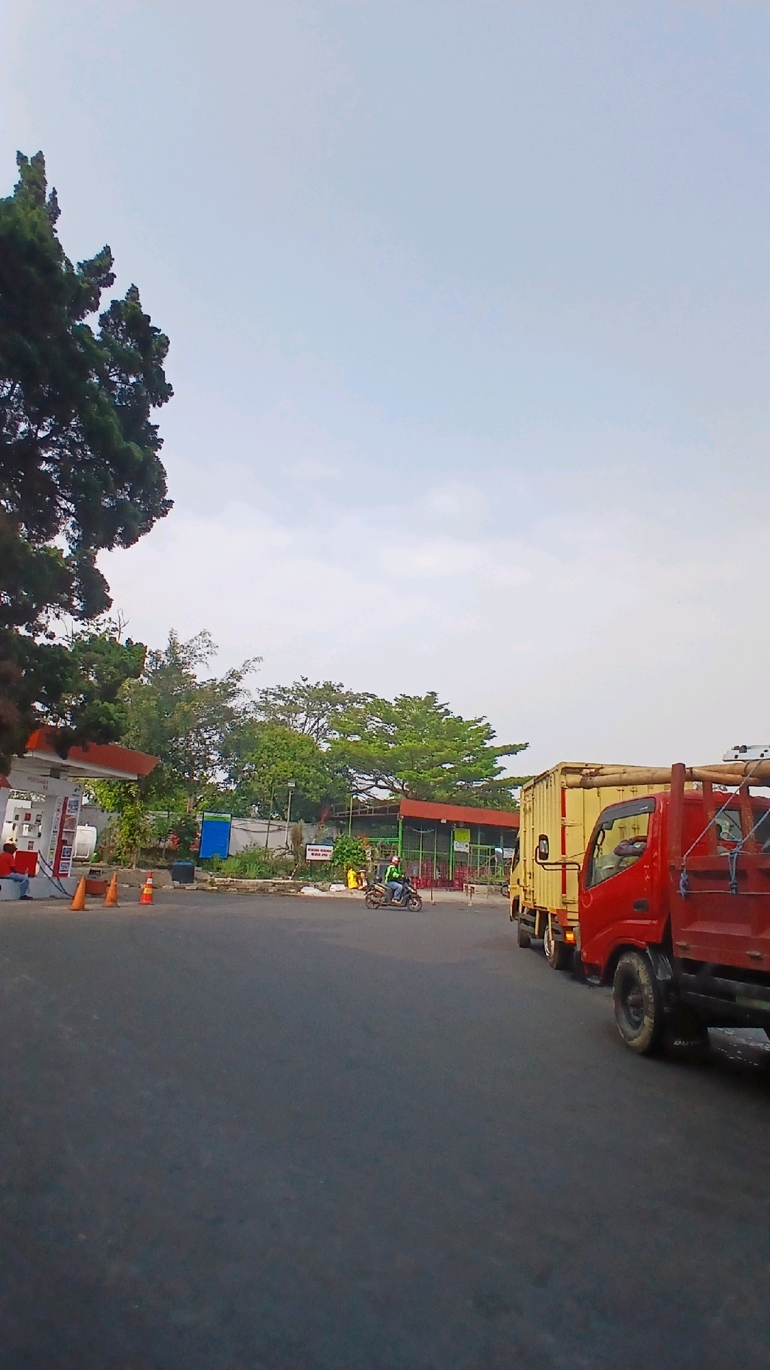The image depicts a view from a vehicle on a street in Bandung City, Indonesia, likely near a gas station. In the foreground, the asphalt road is visible. To the left, a gas station canopy with red trim and white pillars is present, with gas pumps and a person wearing a red shirt visible. Orange traffic cones are placed on the ground. Behind the gas station, a fence separates the area from trees and greenery. A yellow and red truck is parked on the right side of the road. Further down the road, a person on a motorcycle is riding away from the viewer. The sky is overcast and pale blue.