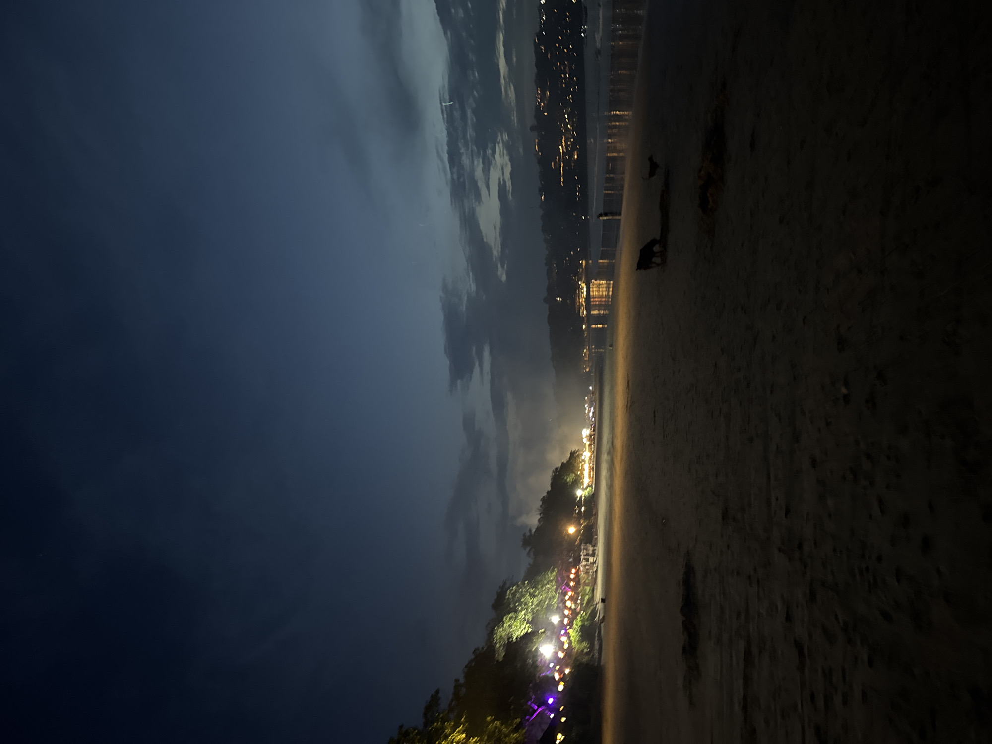 The image depicts a nighttime scene at a beach in Kuta Selatan, Indonesia. The view is oriented vertically, with the dark blue night sky and clouds dominating the left side of the frame. The right side features the sandy beach, dimly lit and appearing dark. There are a few people and possibly animals, silhouettes, present on the beach along the shore. In the middle ground, string lights illuminate the trees and vegetation along the shoreline. Across the water, lights from buildings on the opposite shore are visible, indicating a built-up area. The overall scene appears to be peaceful, capturing the atmosphere of a quiet night on a beach in Kuta Selatan.