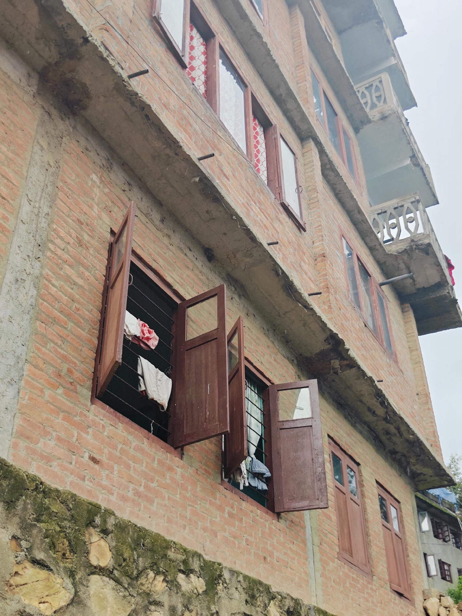 The image depicts a multi-story brick building in Kirtipur, Nepal, with a cloudy sky overhead. The building features several open wooden-shuttered windows, with laundry hanging from a clothesline visible in one of the windows. The lower portion of the building's exterior is a stone wall covered in moss and vegetation. The building's construction appears somewhat unfinished, with exposed concrete and some metal rods protruding from the concrete ledges. In the background, other buildings and structures are visible, suggesting a densely populated area. The perspective is upward, looking at the side of the building from a low angle.