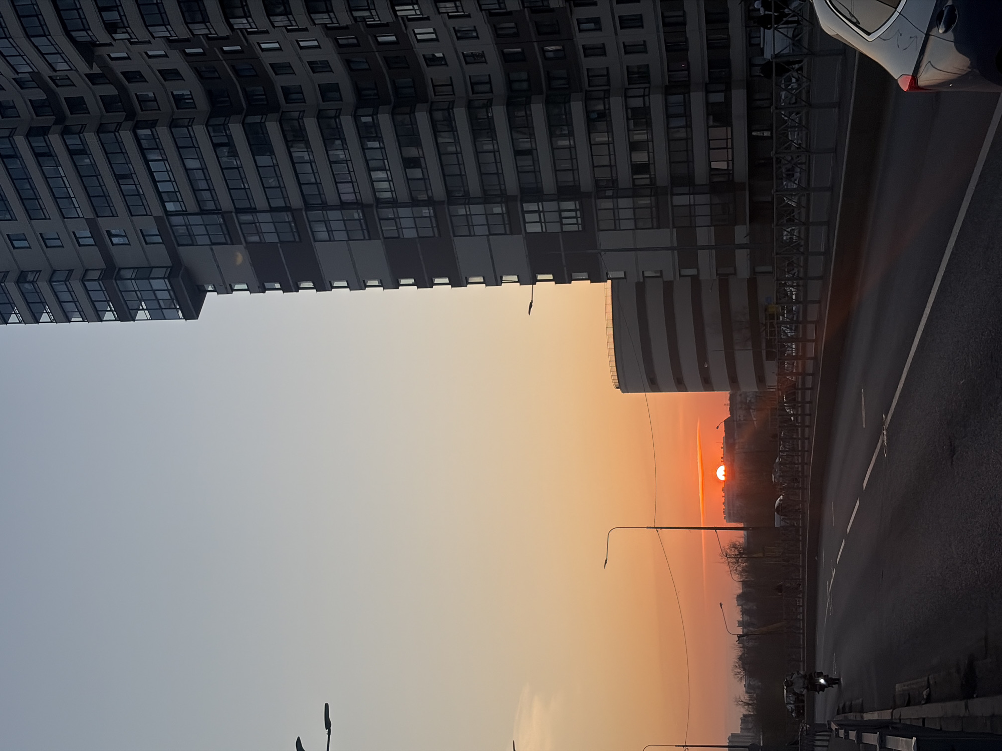 The image shows a sunset in what appears to be an urban setting. A tall residential or office building is on the left side, while a highway or wide road runs along the right. The sun is setting on the horizon, casting an orange glow across the sky. A motorcycle can be seen driving down the highway. Streetlights are visible in the foreground and background. Based on architectural details, this image is likely taken in the city of Saint Petersburg, Russia.