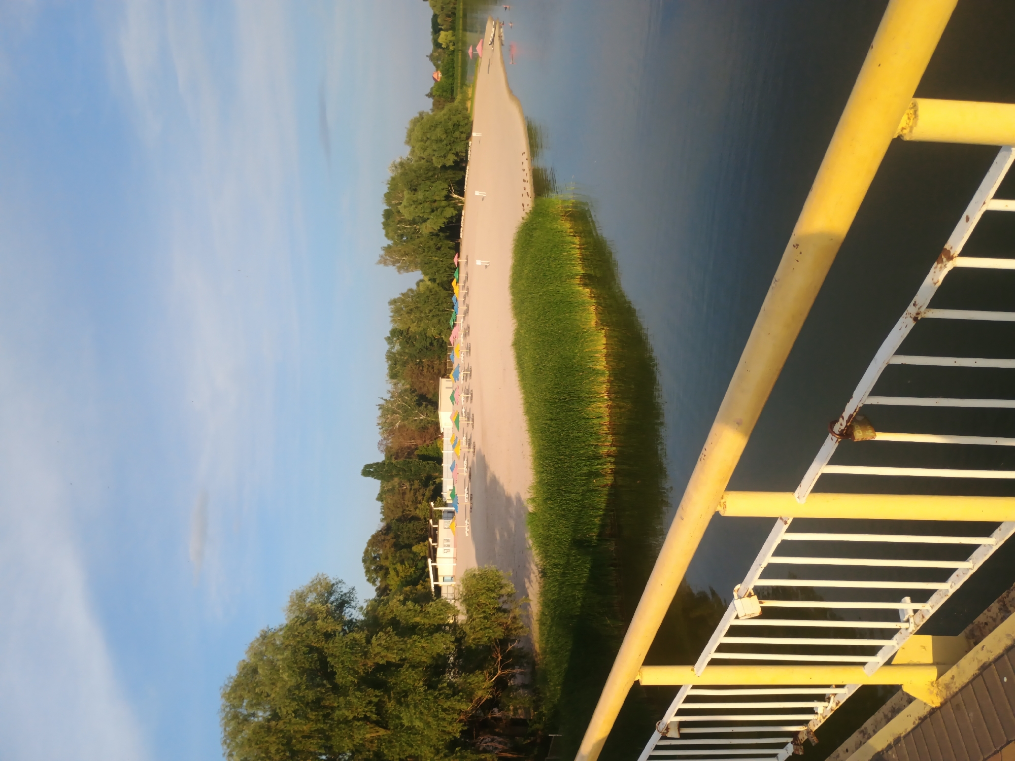 A scenic view of a public beach and lake in Myrhorod, Ukraine, is captured from a bridge. The sun is low in the sky, casting a warm glow on the scene. A sandy beach is lined with colorful umbrellas and lounge chairs, suggesting a recreational area. Dense green trees form a backdrop to the beach, with a few buildings visible further in the distance. The foreground is dominated by the edge of the lake, its calm waters reflecting the sky and surrounding greenery. A yellow-railed bridge, with a white picket fence and some weathered padlocks, extends into the frame, implying the perspective of someone on a walkway overlooking the water and beach. The overall impression is one of tranquility and natural beauty, typical of a lakeside resort.