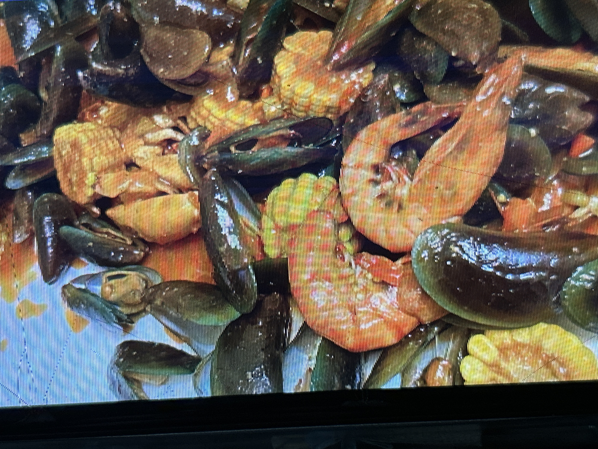 The image displays a close-up, overhead view of a seafood dish, likely a boil, presented on a textured surface with a blue background. The primary components of the dish include mussels and shrimp, interspersed with pieces of corn. The mussels are predominantly dark green to black, with some appearing lighter due to their shells being partially open. The shrimp are a vibrant orange-red, cooked and curled. The corn kernels are a bright yellow. The seafood and corn are coated in a reddish-orange sauce, suggesting a spicy seasoning. The image is captured with a shallow depth of field, focusing on the central elements of the dish. The background is blurred, but appears to be a blue surface, possibly a table or serving platter. The overall presentation is casual and appetizing, typical of a seafood restaurant or home-cooked meal. There are no people or animals visible in the image, and no specific urban or environmental context is provided to indicate a location in Depok, Indonesia. The image quality is affected by visible horizontal lines, suggesting it was captured from a screen display.