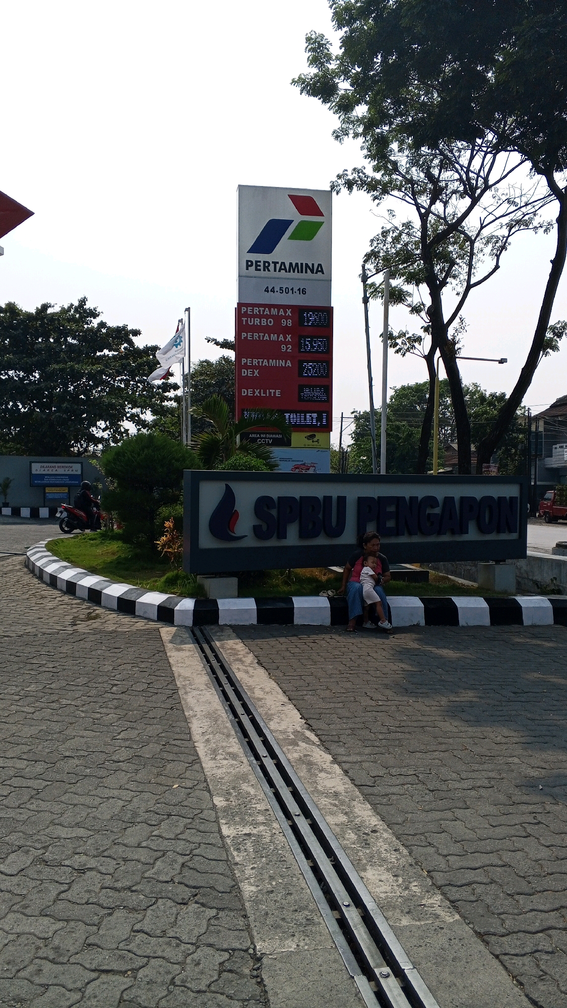 The image depicts a Pertamina gas station in Semarang, Indonesia. A prominent red and white sign with the Pertamina logo displays fuel prices: PERTAMAX TURBO 98 at 13,900, PERTAMAX 92 at 13,500, PERTAMINA DEX at 15,700, and DEXLITE at 15,100. Below this, a large dark gray sign reads "SPBU PENGAFON". A woman and a child are seated on a low concrete barrier in front of the "SPBU PENGAFON" sign. The ground is paved with interlocking gray bricks, and a metal drainage channel runs through the paved area. In the background, trees, a building, and a scooter can be seen. The sky is bright and overcast.