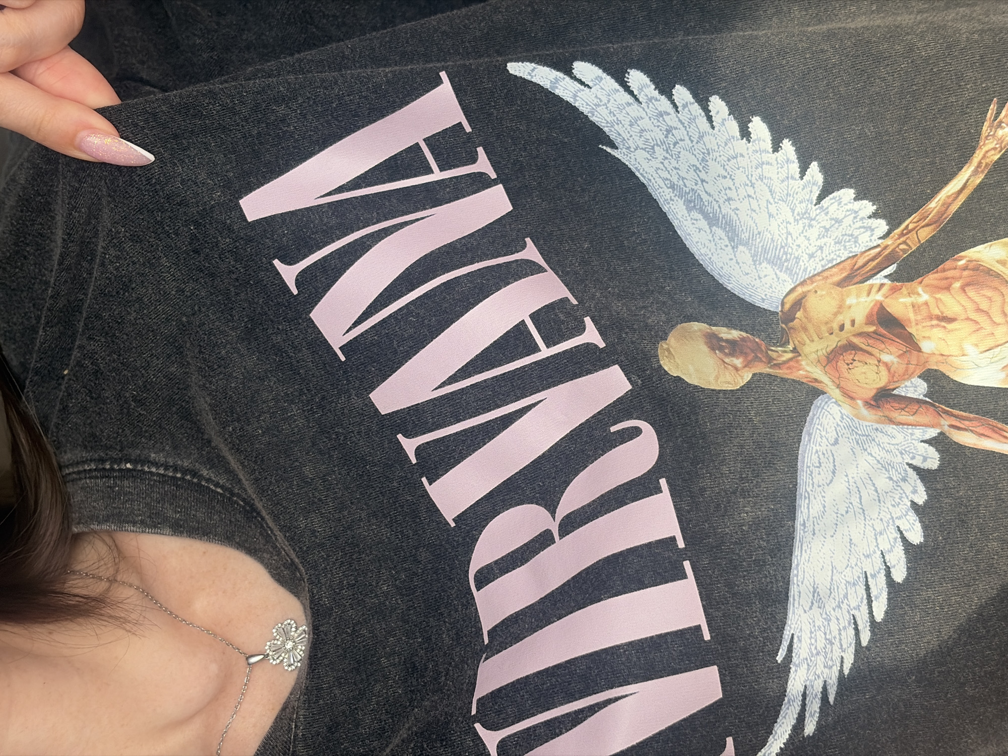A close-up shot displays a black garment featuring a pink "NIRVANA" inscription and a depiction of an anatomical human figure with white wings. A person with manicured nails holds the garment. The person also wears a silver chain necklace with a flower pendant. No information indicates that the scene is in Saint Petersburg, Russia.
