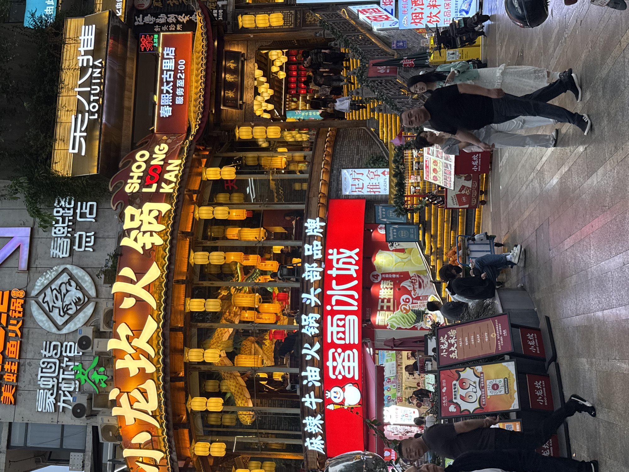 The image shows a street scene in the Jinjiang District of Chengdu, China. Several brightly lit storefronts dominate the view, including a 'SHOO LOONG KAN' hotpot restaurant and a 'LOTUINA' business. A '蜜雪冰城' (Mixue Bingcheng) dessert shop sign is also visible. The area is busy, with people walking along the street. Signs in Chinese advertise various services and products, including hotpot, foot massages, and drinks. A motorcycle is parked on the right side of the image. There are several promotional stands on the ground. The scene is well-lit with numerous lanterns and neon signs.