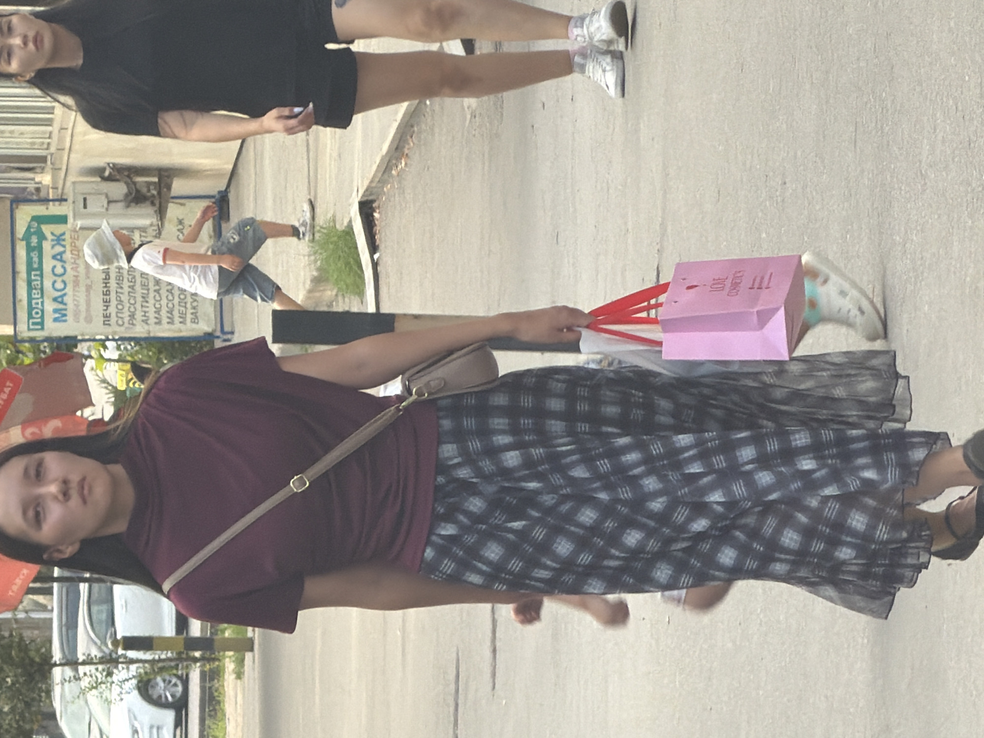 In an urban setting, a young woman with dark hair and a maroon top walks on a sidewalk. She wears a long, dark-and-white plaid skirt, black flats, and a beige crossbody bag. In her right hand, she carries a pink shopping bag with red handles. Behind her, a young child in a light-colored hat, white t-shirt, and blue shorts walks. To the right of the woman, another person in a black top and dark shorts walks. The background shows a white car, a sign with Russian text, and a building. The environment appears to be an outdoor public space with paved surfaces and some vegetation.
