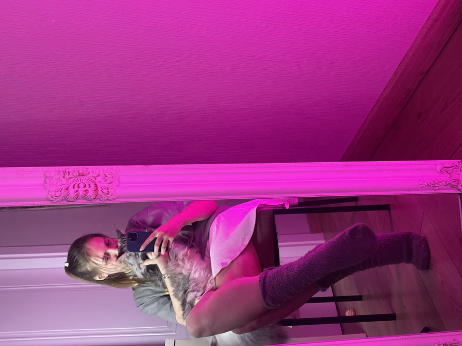 In a mirror reflection bathed in pink light, a young woman wearing gray clothing and fuzzy gray socks sits with crossed legs, holding a gray cat close to her face. She's taking a photo of the cat with her phone. The room features a light-colored wall and wooden paneling. A chair is visible in the background of the mirror. The mirror has a white, ornate frame.