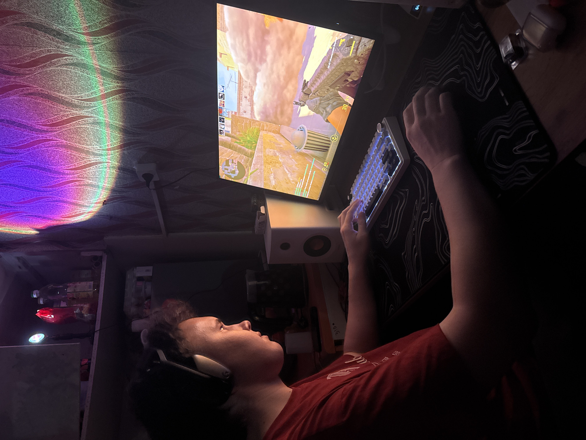 A person with headphones is playing a video game on a computer. The game appears to be a first-person shooter, judging by the weapon visible on the screen. The person is using a keyboard and mouse. The room is dimly lit with a projector light casting a rainbow effect on the wall. There is also a speaker and a shelf with items on it. It is not possible to determine the specific location in Perm, Russia, or confirm this location at all without additional evidence.