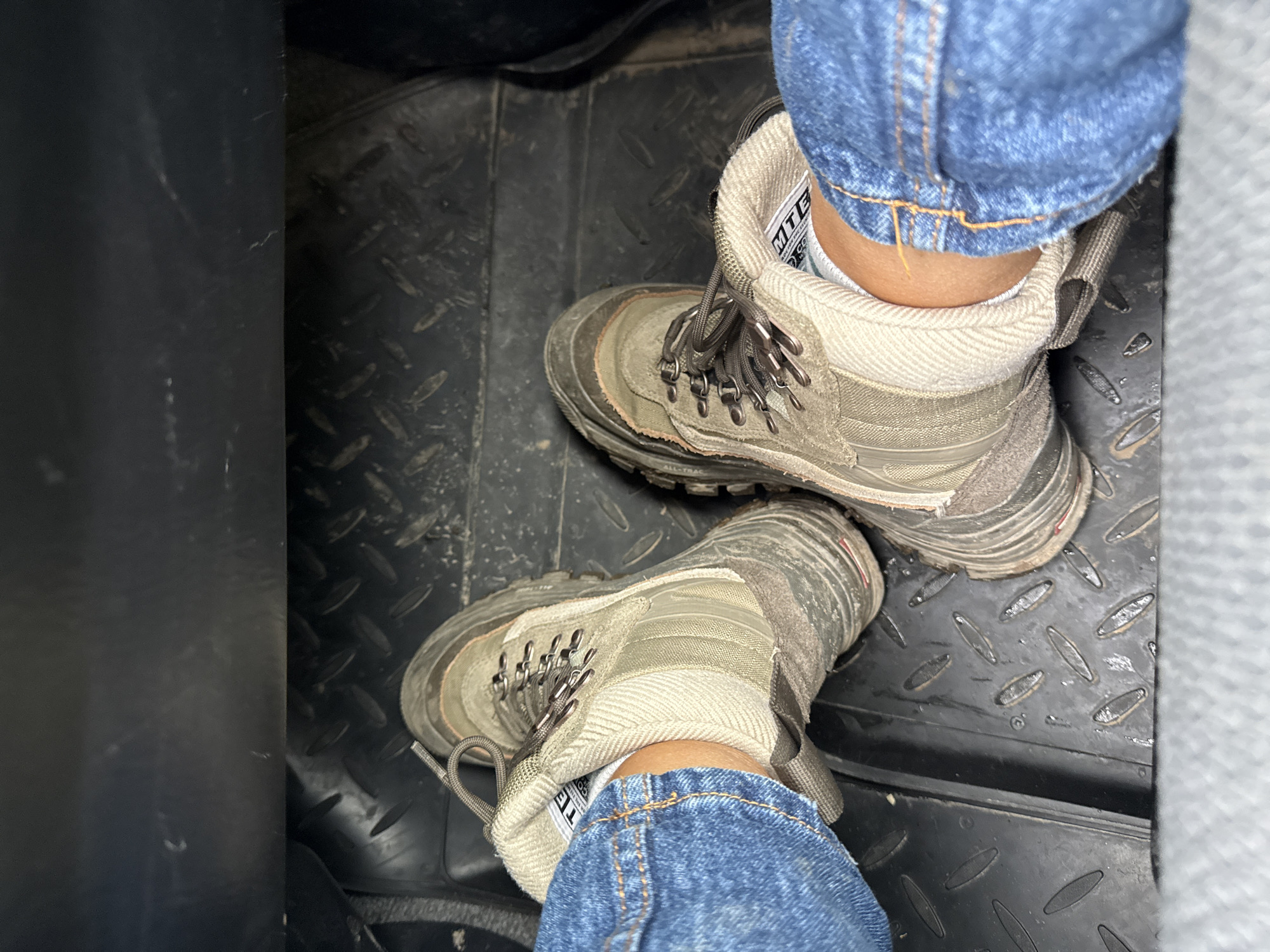 The image shows a pair of worn hiking boots on a black rubber mat with a diamond pattern. The boots are laced and appear to be made of a light-colored canvas material with darker trim. A person wearing blue jeans has their feet in the boots. The setting appears to be the floor of a vehicle. There is no information available to determine if the location is Perm, Russia.
