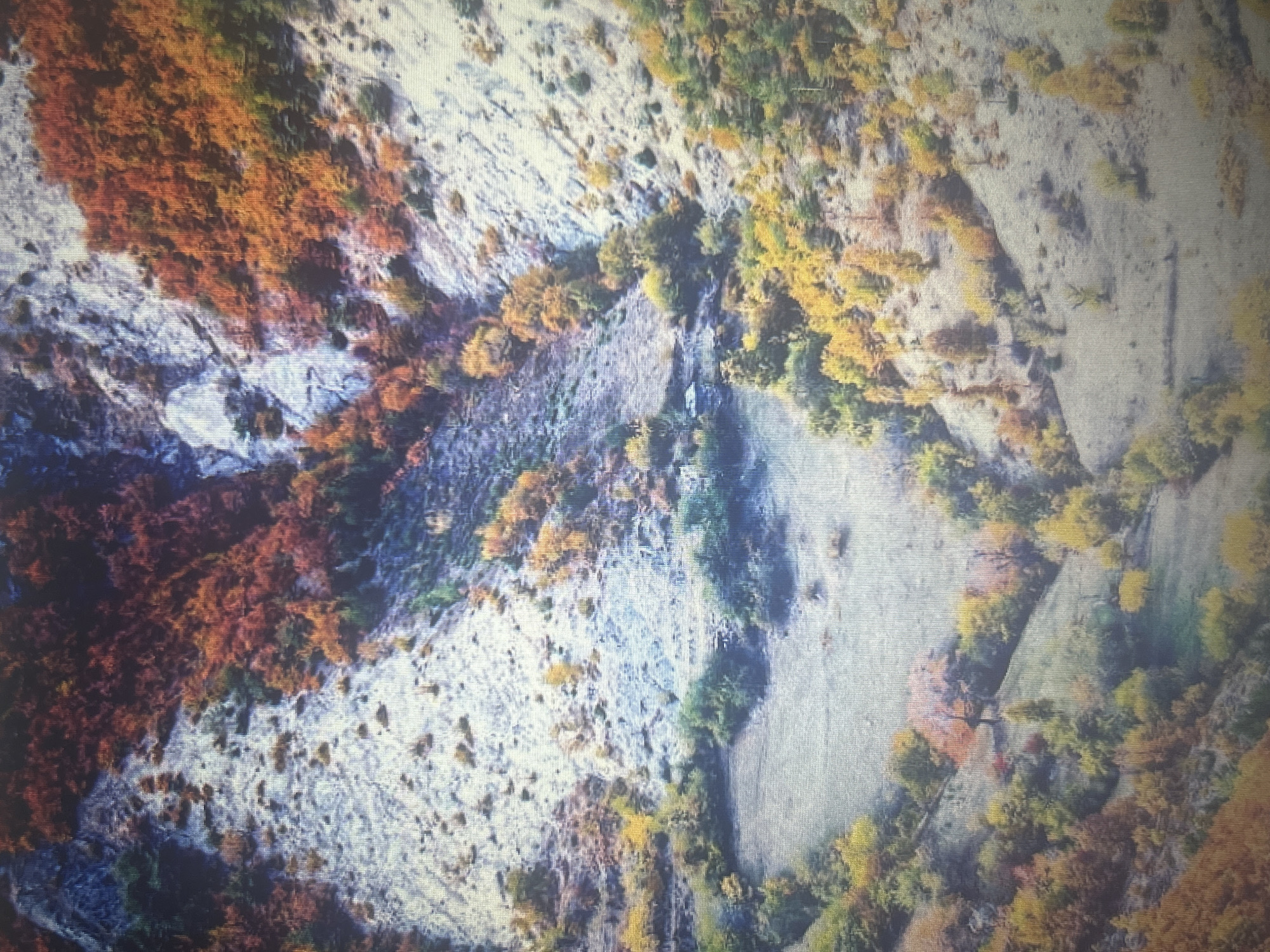 The image depicts an aerial view of a mountainous landscape during autumn. The terrain is characterized by steep, rocky slopes covered in a mix of deciduous trees displaying vibrant fall foliage in shades of red, orange, and yellow. A small, cleared area resembling a field or meadow is visible in the lower half of the image, bordered by trees. The scene suggests a natural, possibly rural, environment. There are no people, animals, or man-made structures visible in the image. The image is a photograph of a natural landscape in an undisclosed location. There is no information to suggest this location is in the city of Sofia, Bulgaria.