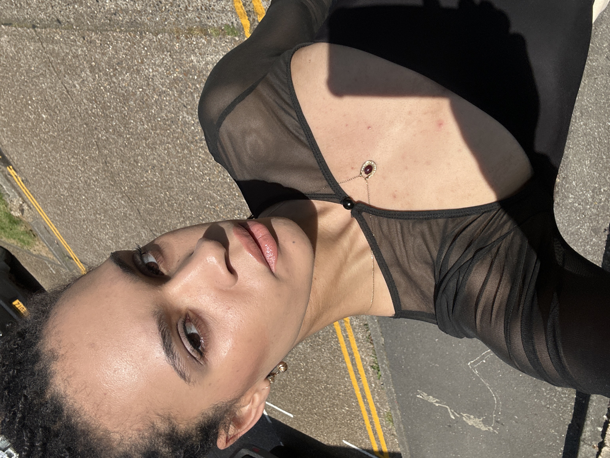 The image shows a person with brown skin and curly dark hair wearing a black sheer top with a teardrop-shaped pendant necklace. The background shows an asphalt surface with yellow lines marking lanes. The location appears to be a street in London, United Kingdom.