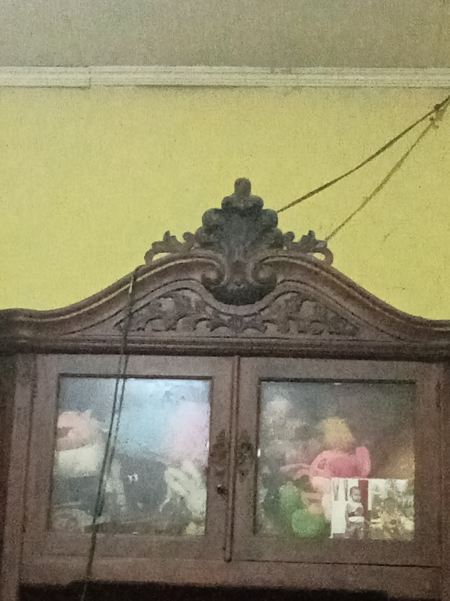 The image shows the upper portion of an ornate wooden cabinet with glass doors. The cabinet has intricate carvings on its crest and frame. Inside the cabinet, visible through the glass, are various objects, including what appear to be stuffed animals and possibly photographs. A dark cord hangs vertically in front of the left glass door. The background is a plain yellow wall with a white molding strip near the ceiling. The lighting is dim, creating shadows and a somewhat muted color palette. The image is framed from a mid-level perspective, focusing on the cabinet and the wall behind it. There are no discernible features that would indicate a specific location in Grabag, Indonesia.