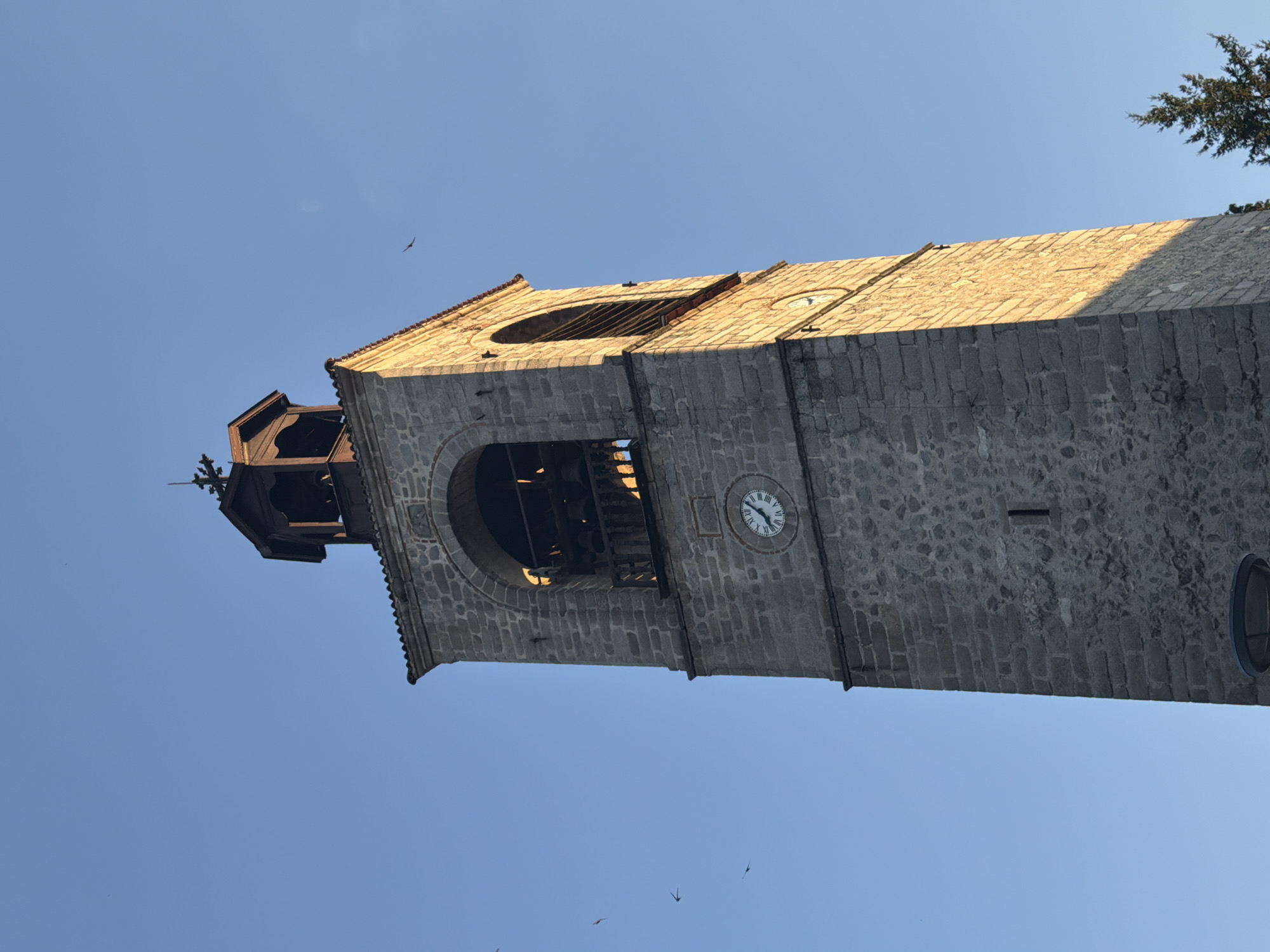 A stone clock tower stands against a clear blue sky. The tower, constructed of rough-hewn stone blocks, has an arched opening near its top, revealing bells and wooden structures within. Below this opening, a round clock face displays Roman numerals. The tower is illuminated by sunlight on its right side, creating a strong shadow on its left. Several birds are visible in flight against the sky. The tower is located in the city of Bansko, Bulgaria.
