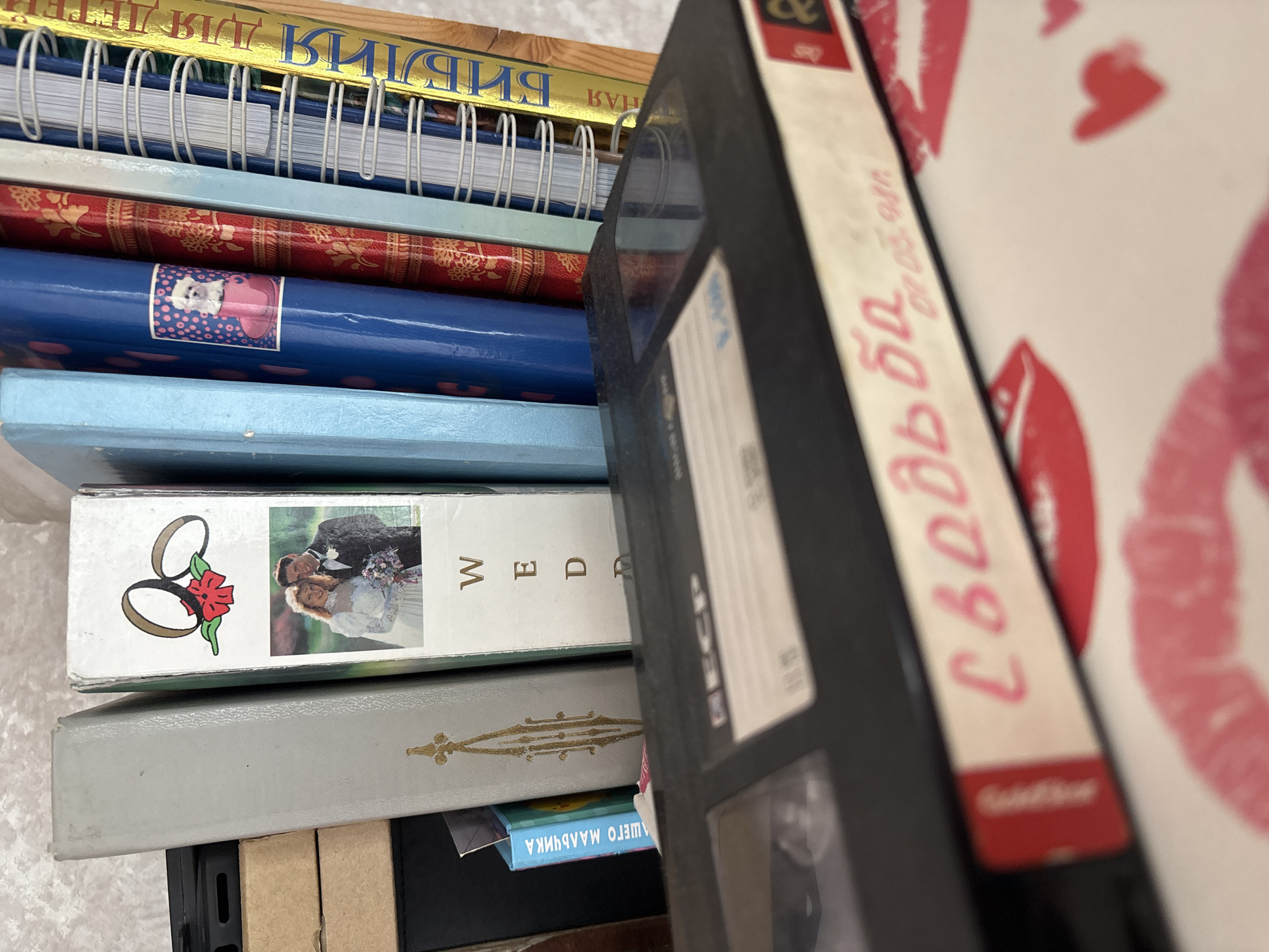 A collection of books and a VHS tape are arranged on a surface. The books are of various colors and sizes, with some featuring decorative designs and text in Cyrillic script. One book has a wedding image on it. The VHS tape in the foreground has a handwritten label with Cyrillic text in pink marker. The surface beneath the VHS tape is decorated with pink lipstick kiss marks and hearts. The lighting is moderate, casting soft shadows. There are no visible people in the image. The scene appears to be indoors, on a shelf or table.