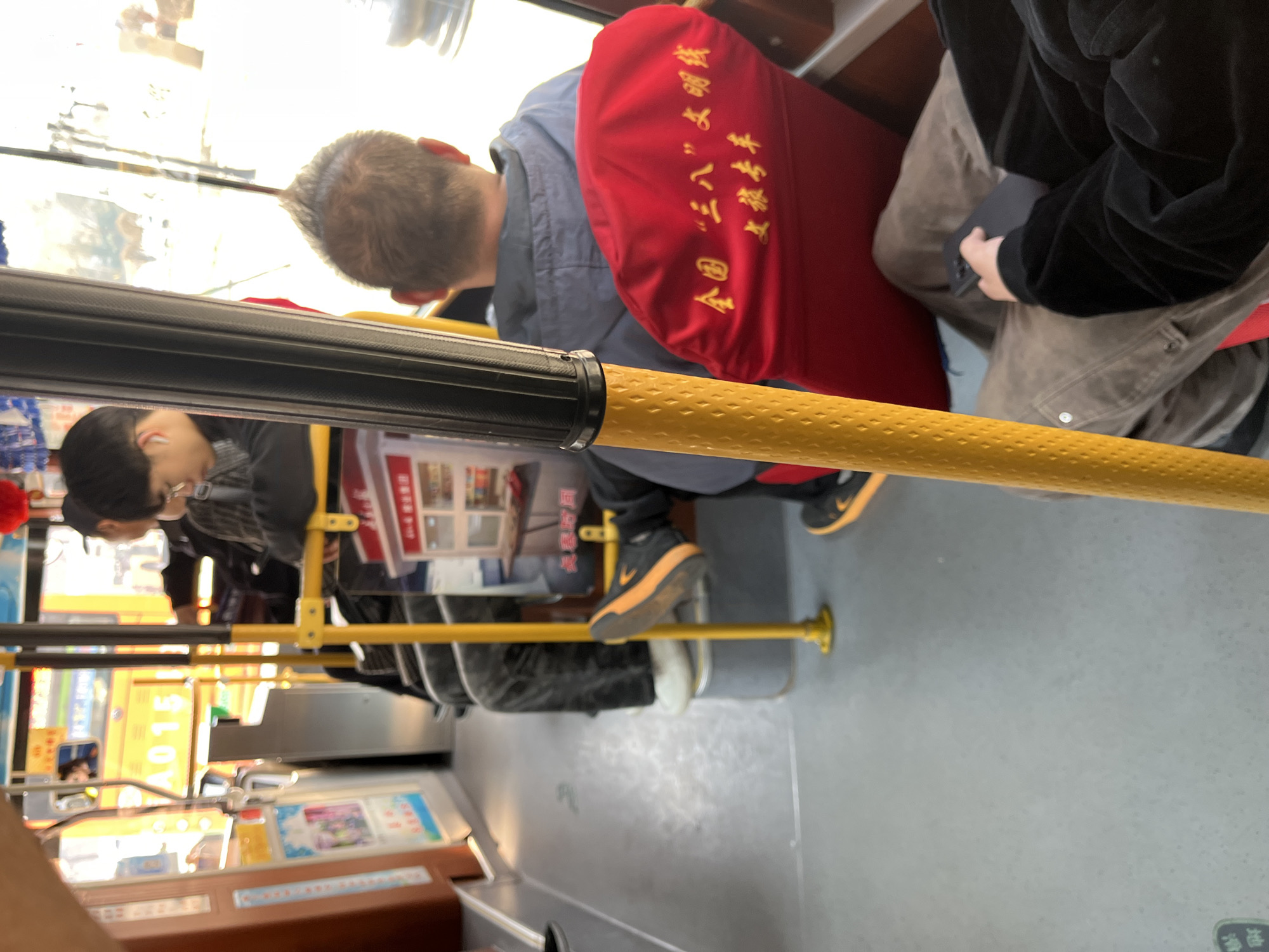 A slightly canted interior shot of a public bus in Changchun City, China. Passengers are seated. A man in a blue jacket sits on a red-cushioned seat adorned with yellow Chinese writing. To his right, another individual in a black velvet jacket is seated, their lap occupied with a dark object. A yellow grab bar runs across the foreground. Visible through the bus window is a scene of the city, with various signs and lights. Other passengers and seats are seen in the background.