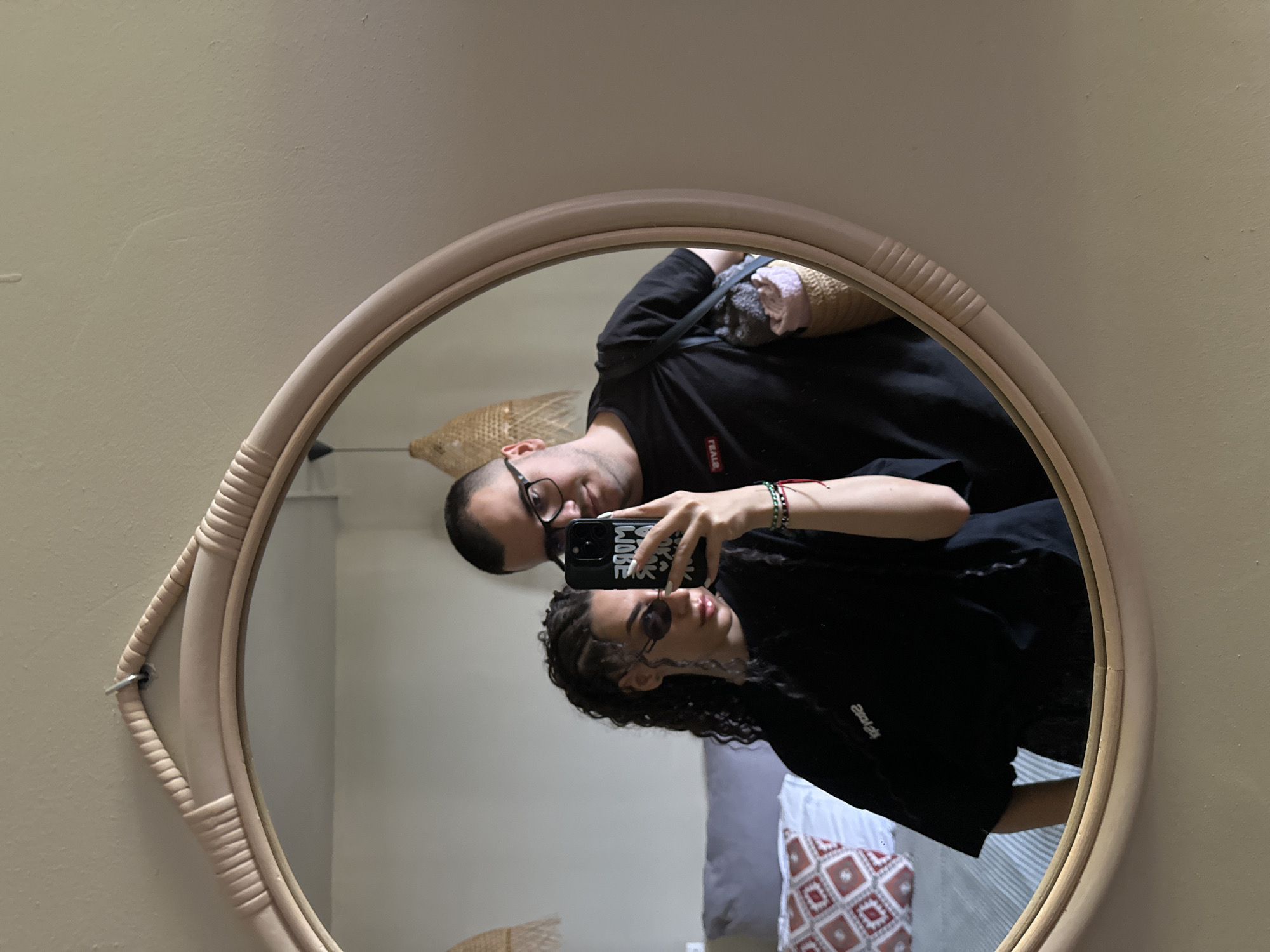 A couple, a woman with braids and sunglasses and a man wearing glasses, take a mirror selfie in a room. The woman is holding a smartphone and looking at the camera, while the man stands behind her, smiling. They are both wearing black t-shirts. In the background, a bed with patterned pillows is visible, suggesting the location is a hotel room. The mirror has a woven, light-colored frame. The image appears to be taken in a residential or hotel setting. The text visible on the woman's shirt, "STAY POSITIVE," and on the man's shirt, "FEAR," suggest a motivational or lifestyle theme. The overall lighting is soft and natural.